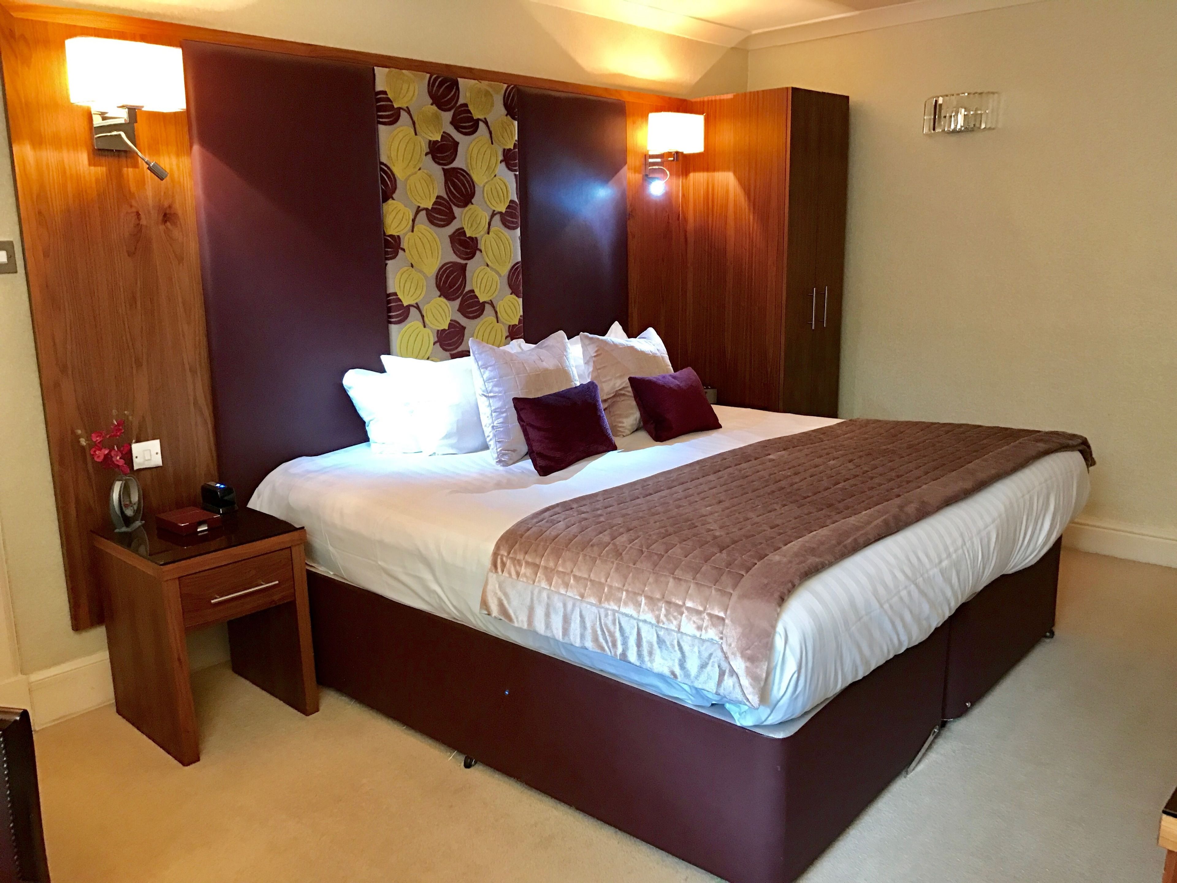 A hotel room with a large bed, wooden headboard, bedside tables, and a wall-mounted lamp.