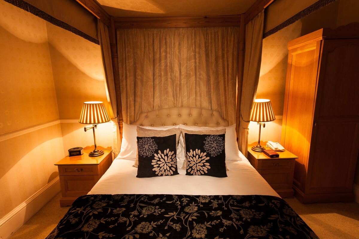 A hotel room with a large bed, two bedside tables with lamps, a wooden wardrobe, and a curtain-covered window.