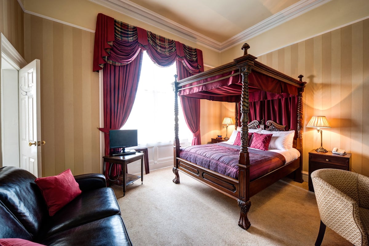 A four-poster bed with a canopy, a leather sofa, a chair, a bedside table, a lamp, and a television.