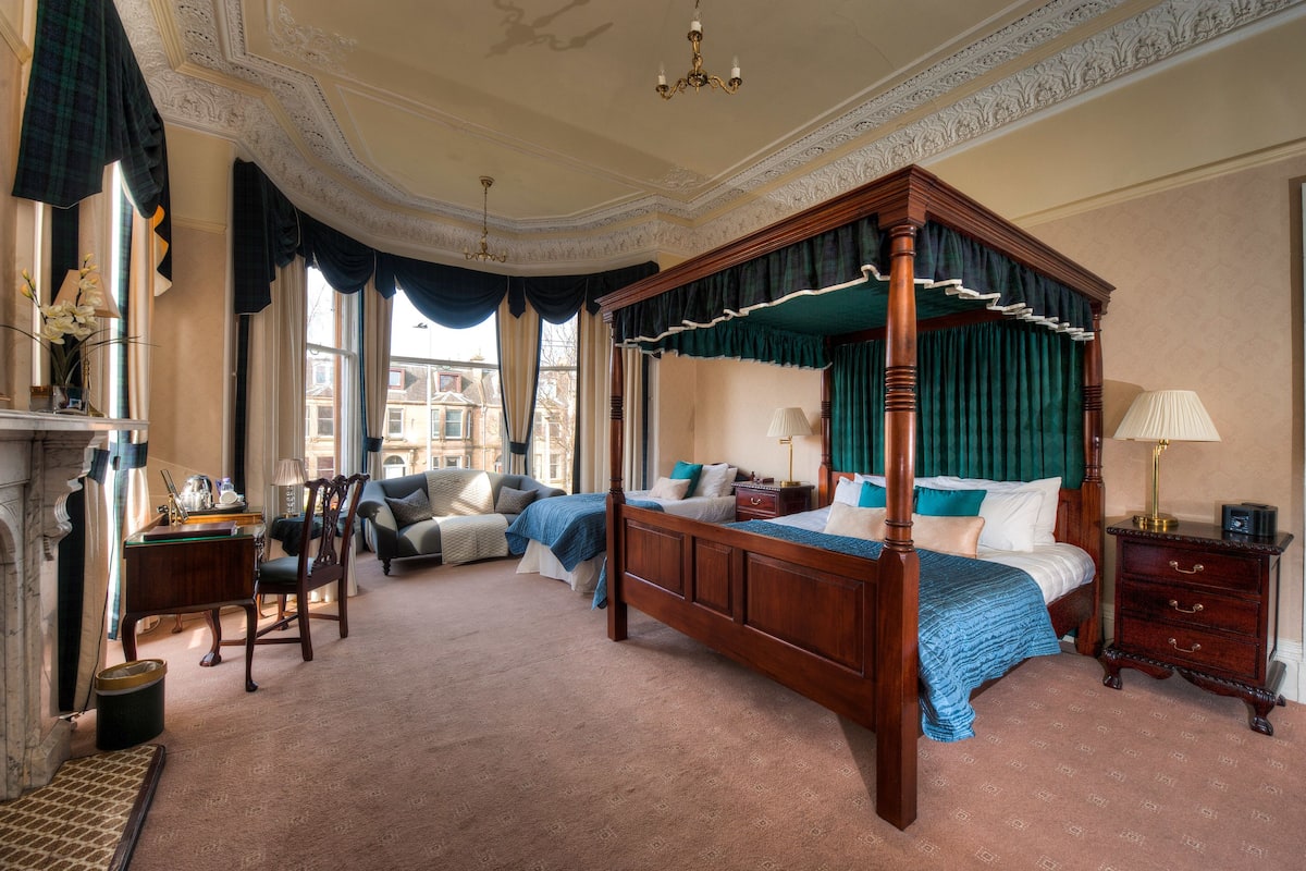 A four-poster bed with a canopy, a sitting area with a sofa and armchairs, a desk with a lamp, and a nightstand with a lamp.