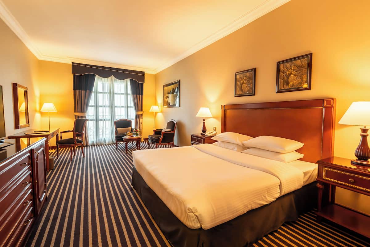 Une chambre d’hôtel avec un grand lit, un bureau, une chaise, une lampe et une fenêtre avec des rideaux.