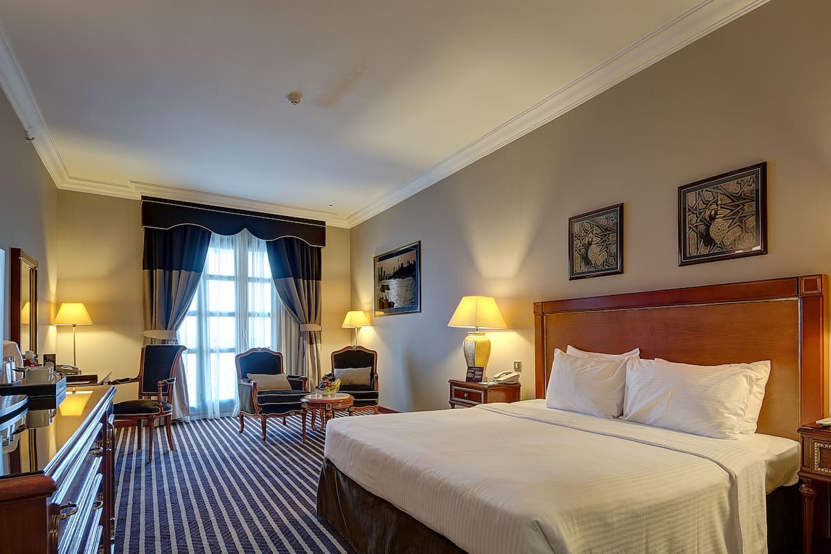Une chambre d’hôtel avec un grand lit, deux tables de chevet, un bureau avec une chaise, une petite table avec une chaise et une fenêtre avec des rideaux.