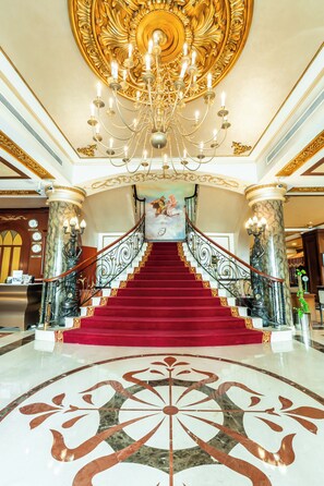 A grand staircase with a red carpet, a chandelier, and ornate columns. - Royal Ascot Hotel (Dubai)