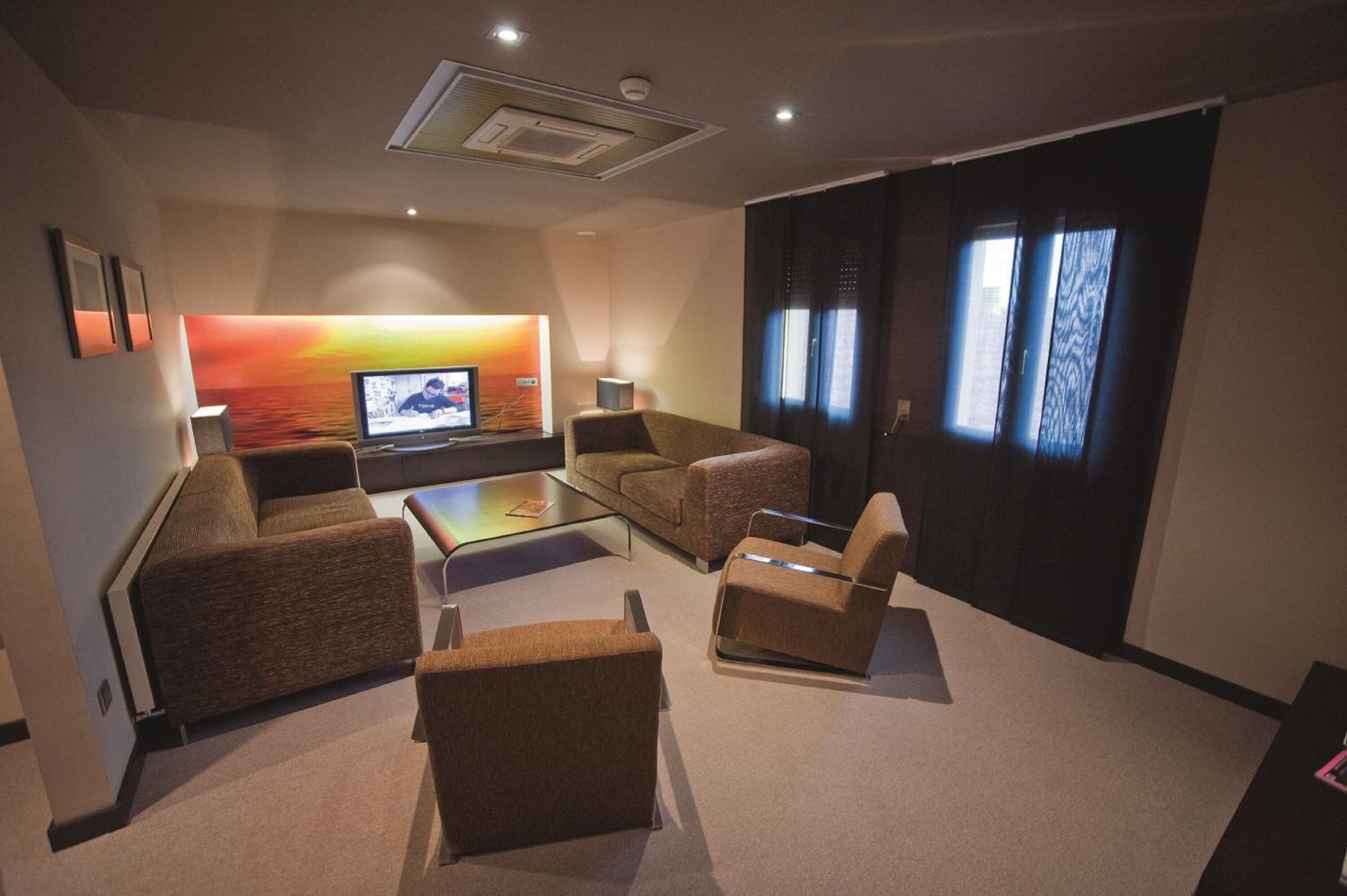 Una sala de estar moderna con televisor de pantalla plana, asientos cómodos y un amplio ventanal con cortinas.