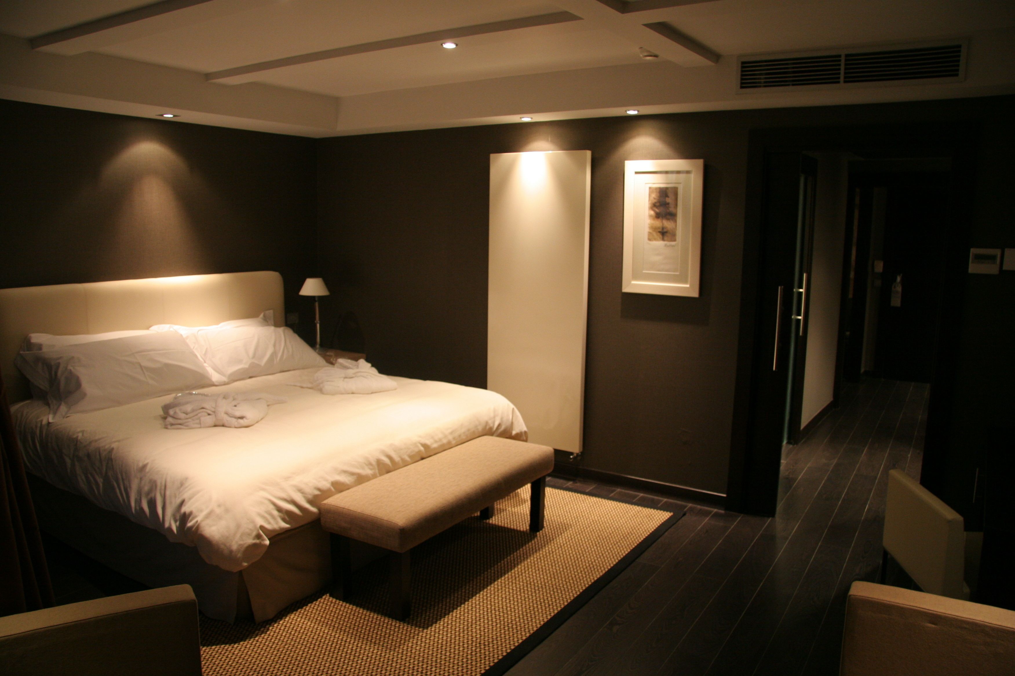 Quarto de hotel com uma cama grande, um banco, uma mesa de cabeceira, uma luminária e um quadro na parede.
