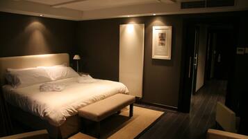 Quarto de hotel com uma cama grande, um banco, uma mesa de cabeceira, uma luminária e um quadro na parede.