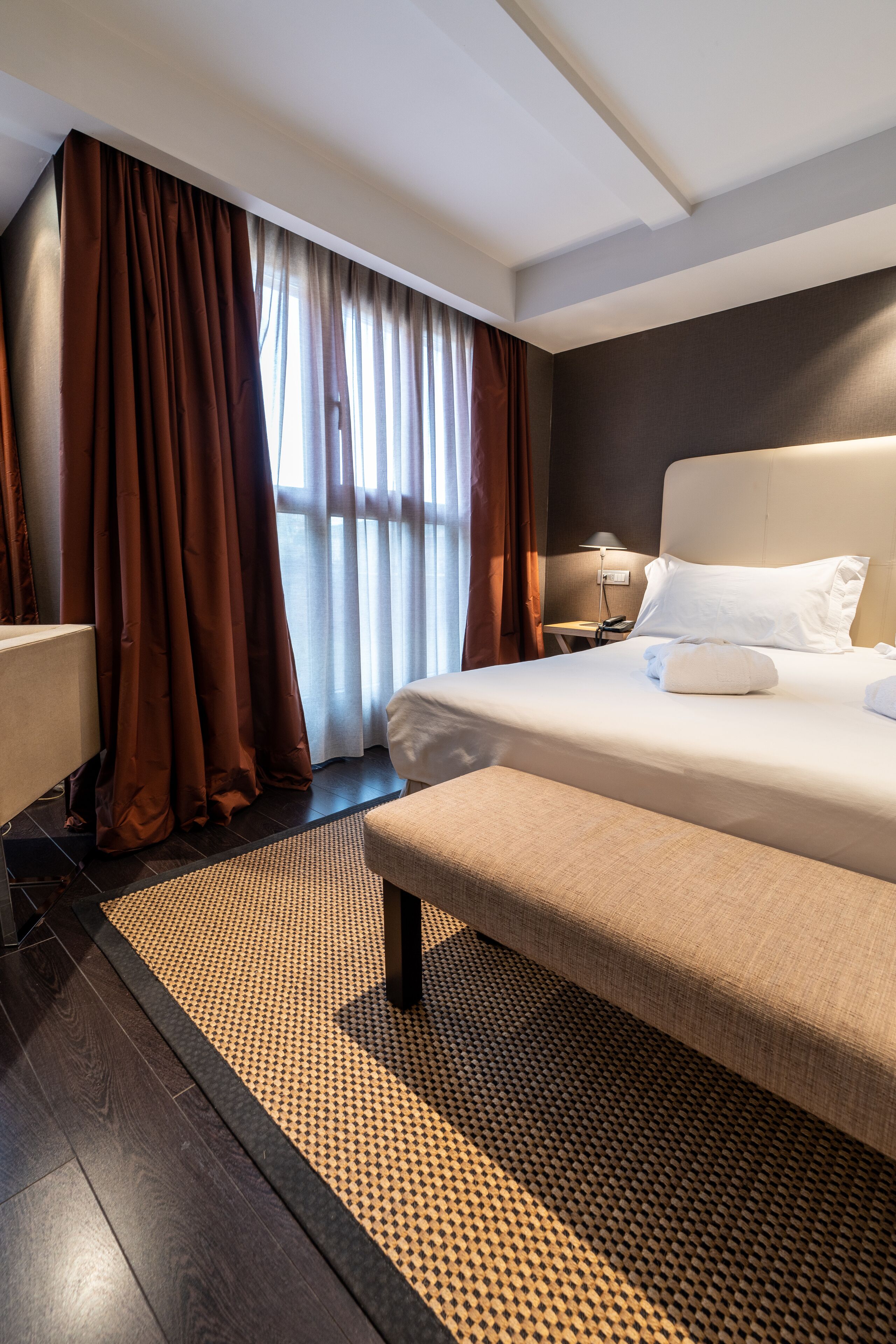 A hotel room with a large bed, a bedside table, a lamp, and a view of the outdoors through the window.