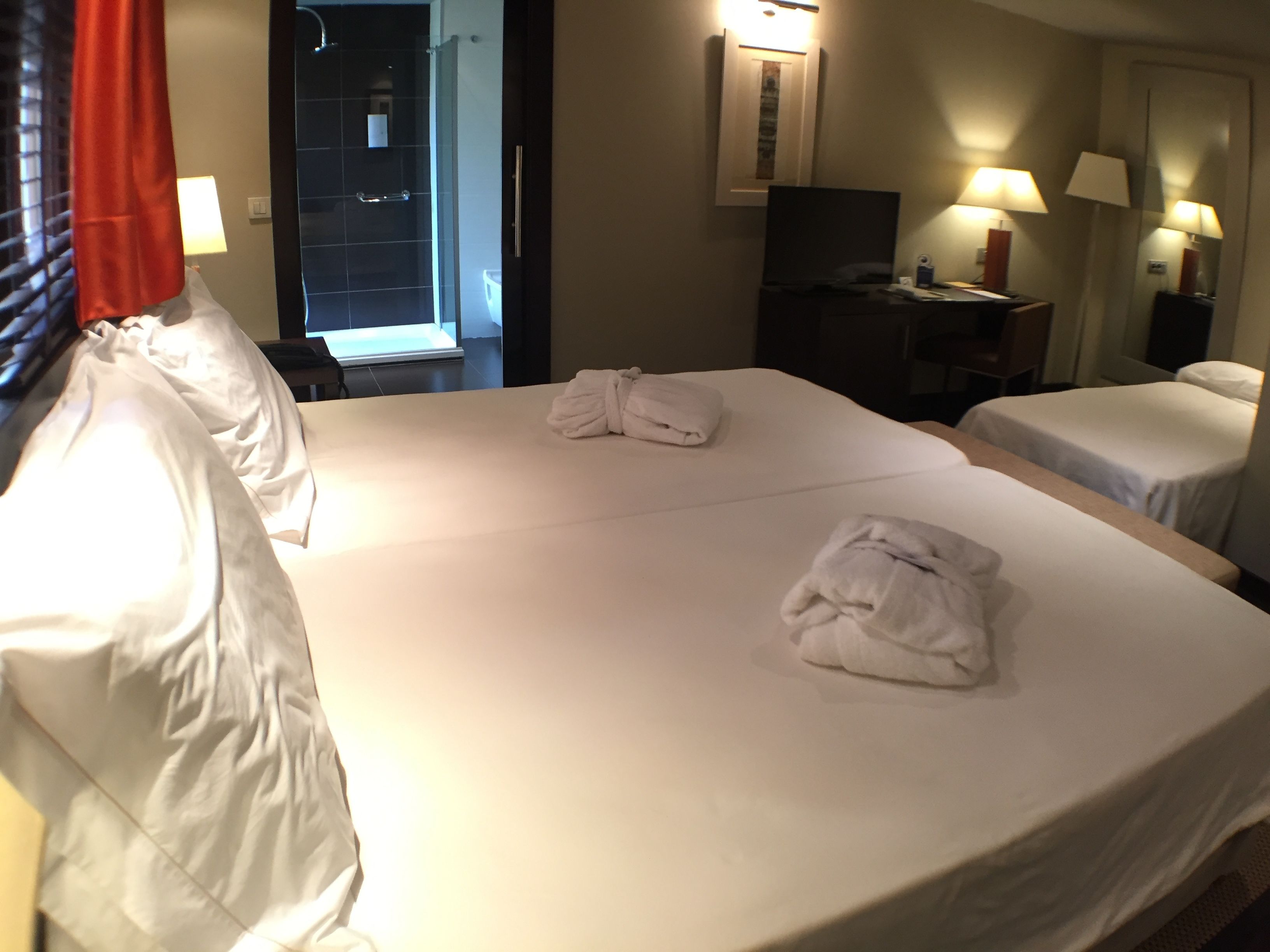 Habitación de hotel con dos camas, un escritorio con televisor y baño visible a través de una puerta abierta.