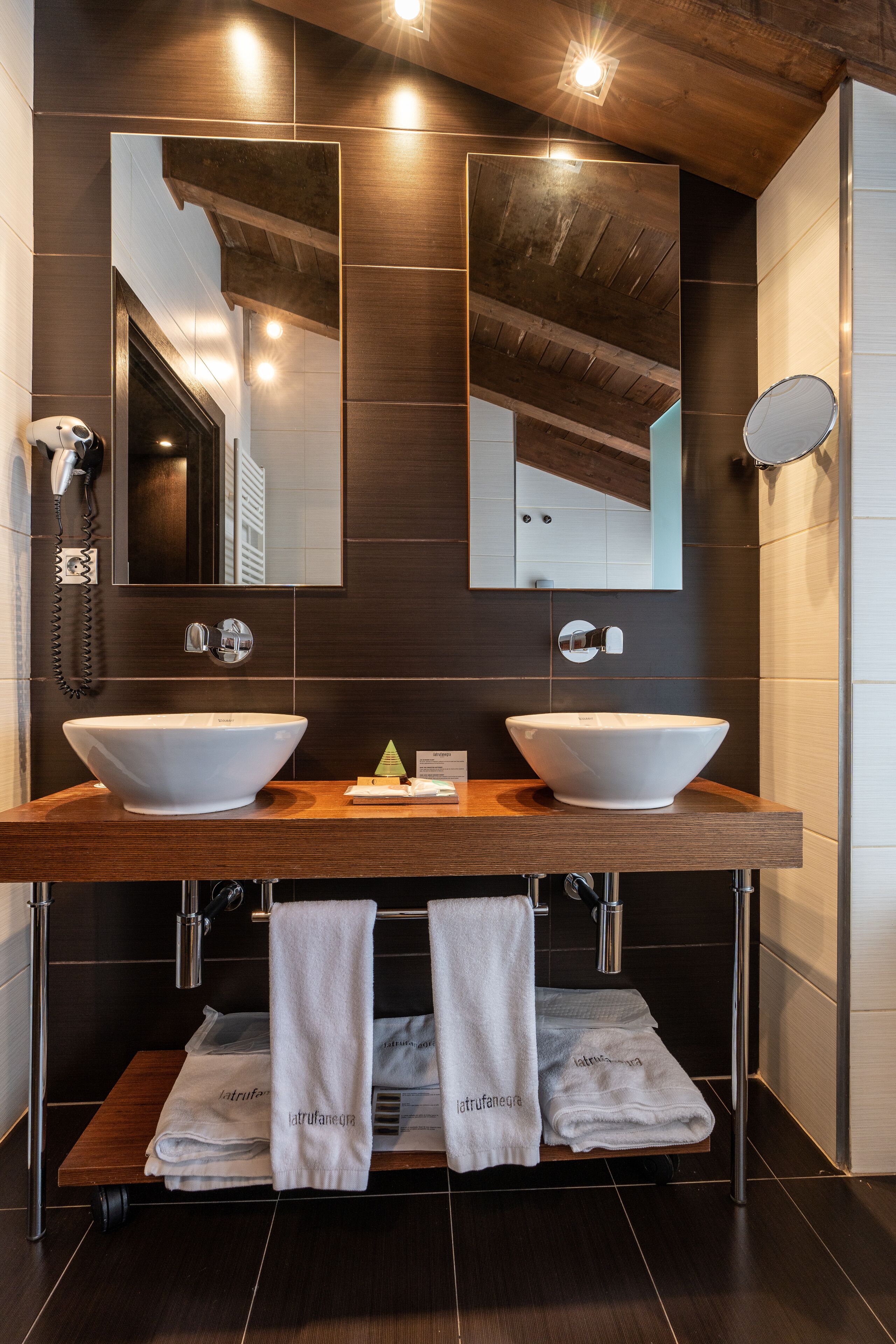 Ein modernes Badezimmer mit zwei großen, weißen Waschtüben, einem Holzauszug und einem großen Spiegel über den Waschbecken. Auf dem Auszug liegen Handtücher und ein kleines, grünes Dekoelement.