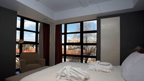 Een hotelkamer met een groot bed, twee ramen met gordijnen, een stoel en uitzicht op gebouwen.