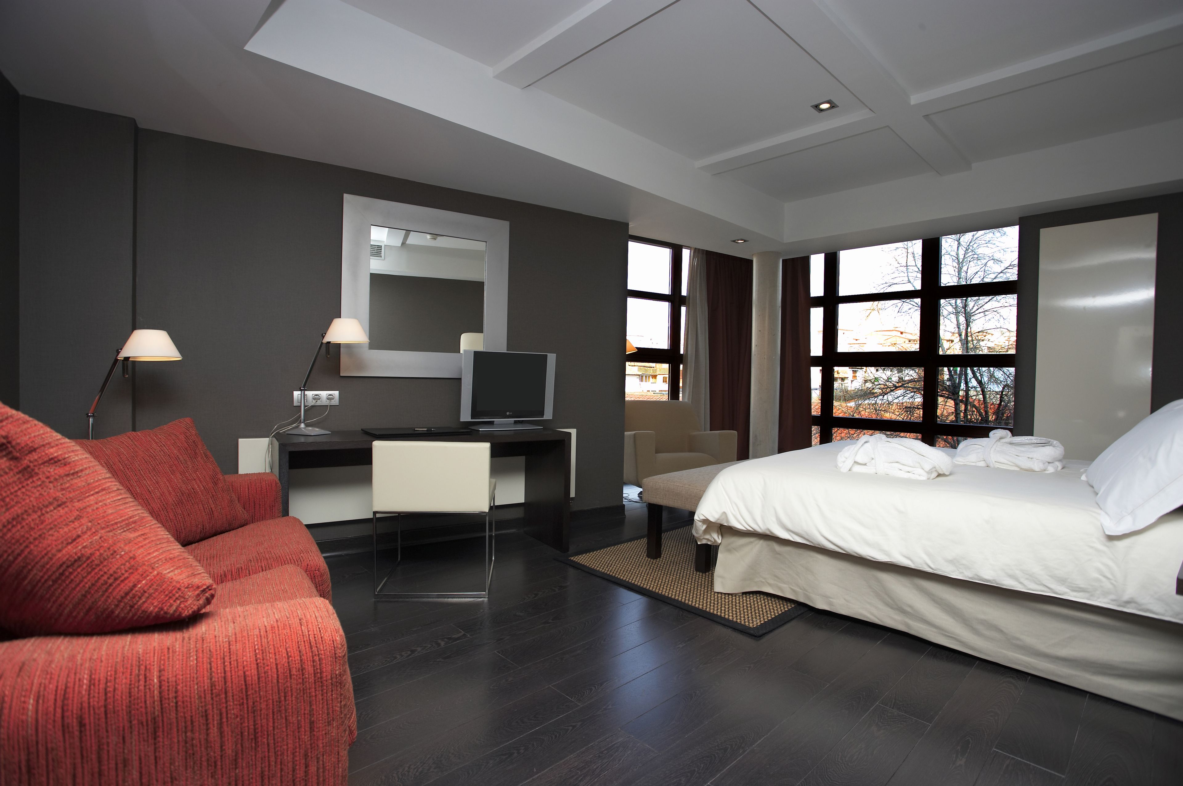 A modern hotel room with a large bed, a desk with a computer, a sofa, and a large window with a view of trees.