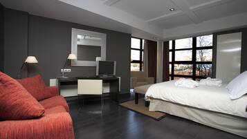 A modern hotel room with a large bed, a desk with a computer, a sofa, and a large window with a view of trees.