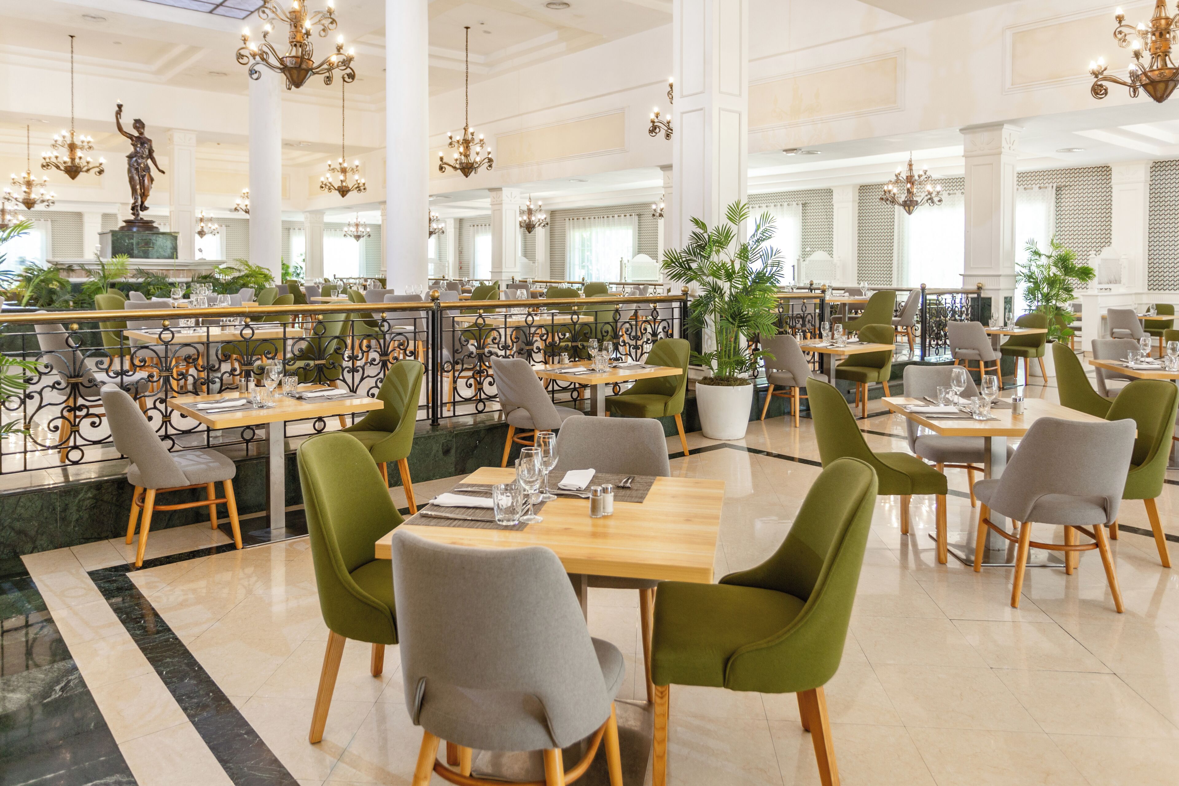 A spacious restaurant with high ceilings, chandeliers, and a statue centerpiece. Tables are set with chairs, and potted plants are placed around the room.