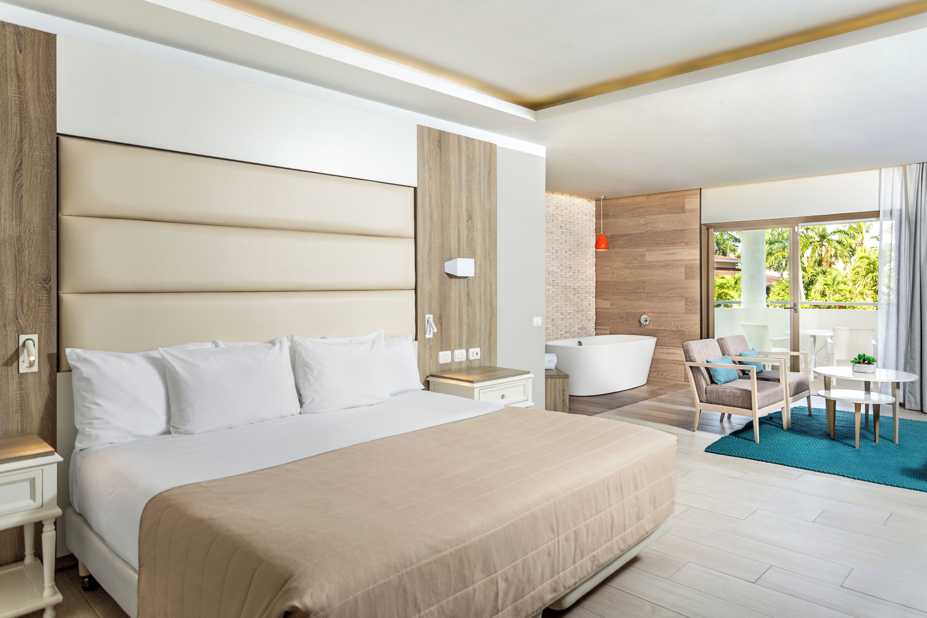 A hotel room with a large bed, a sitting area with a table and chairs, a bathtub, and a view of greenery outside.