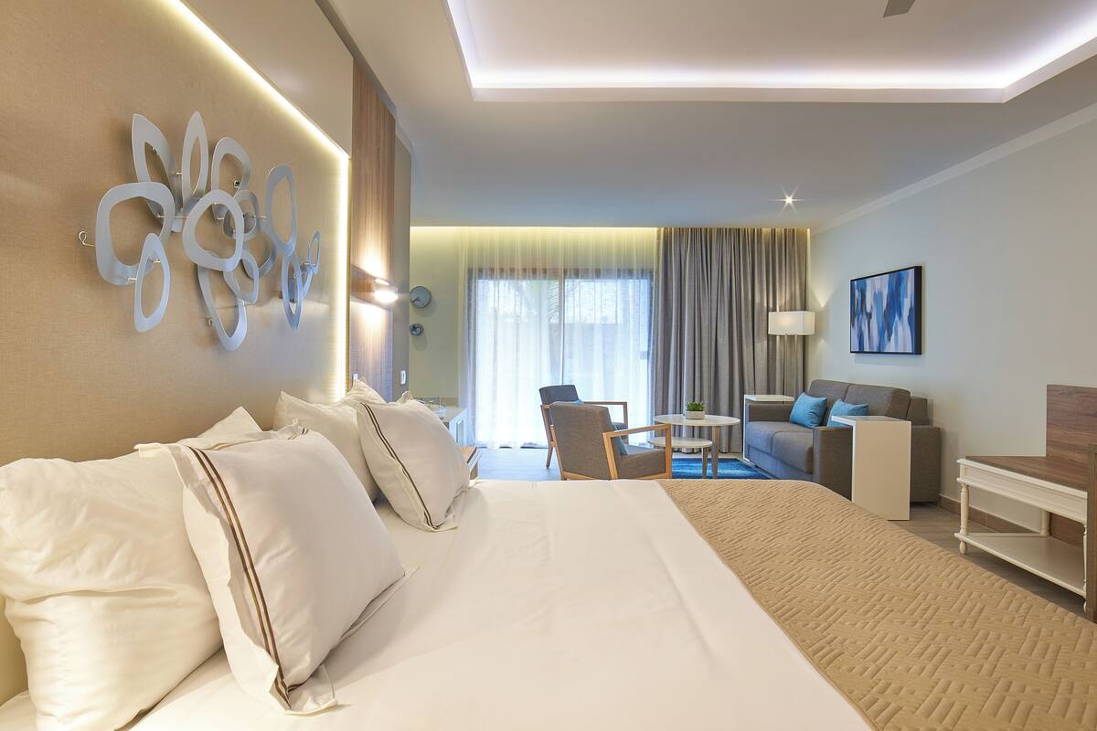 A hotel room with a large bed, a desk, a chair, and a sofa. There is a window with curtains and a painting on the wall.