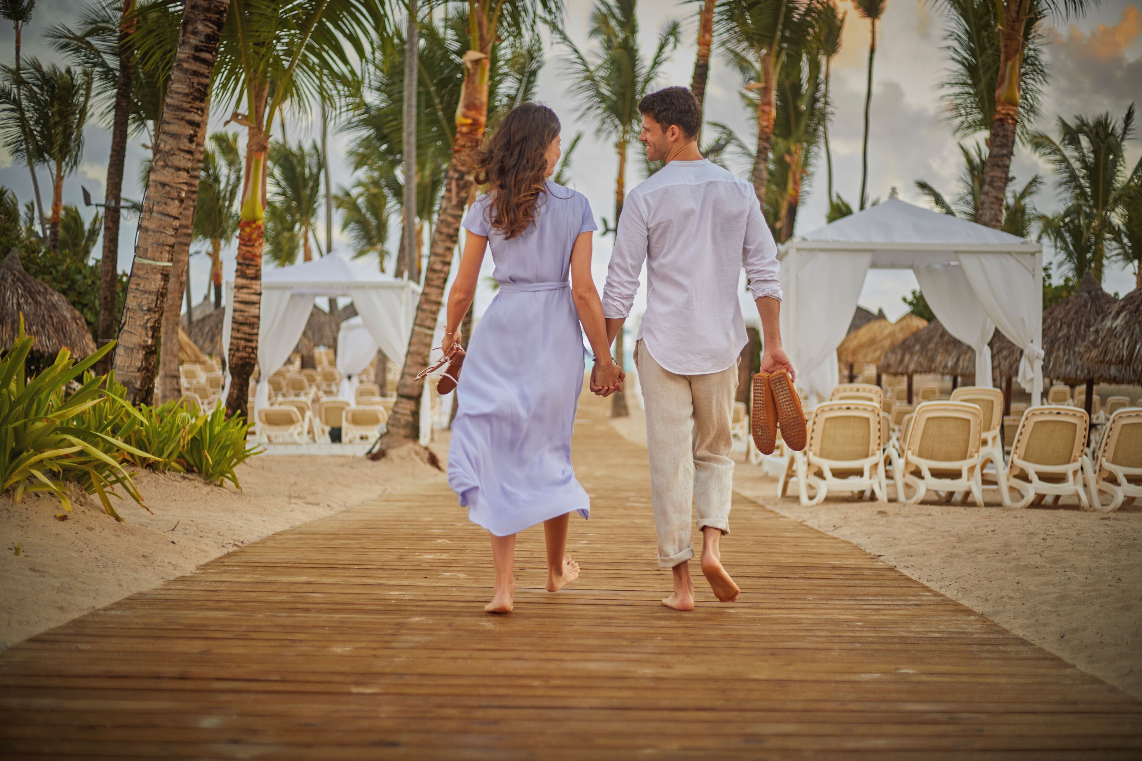 A wooden boardwalk leading to beach chairs and a tent, surrounded by palm trees.