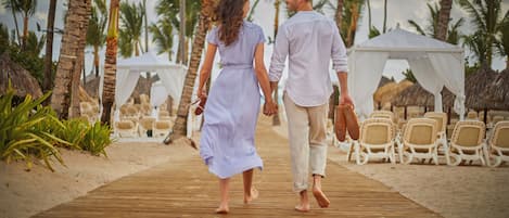 A wooden boardwalk leading to beach chairs and a tent, surrounded by palm trees.