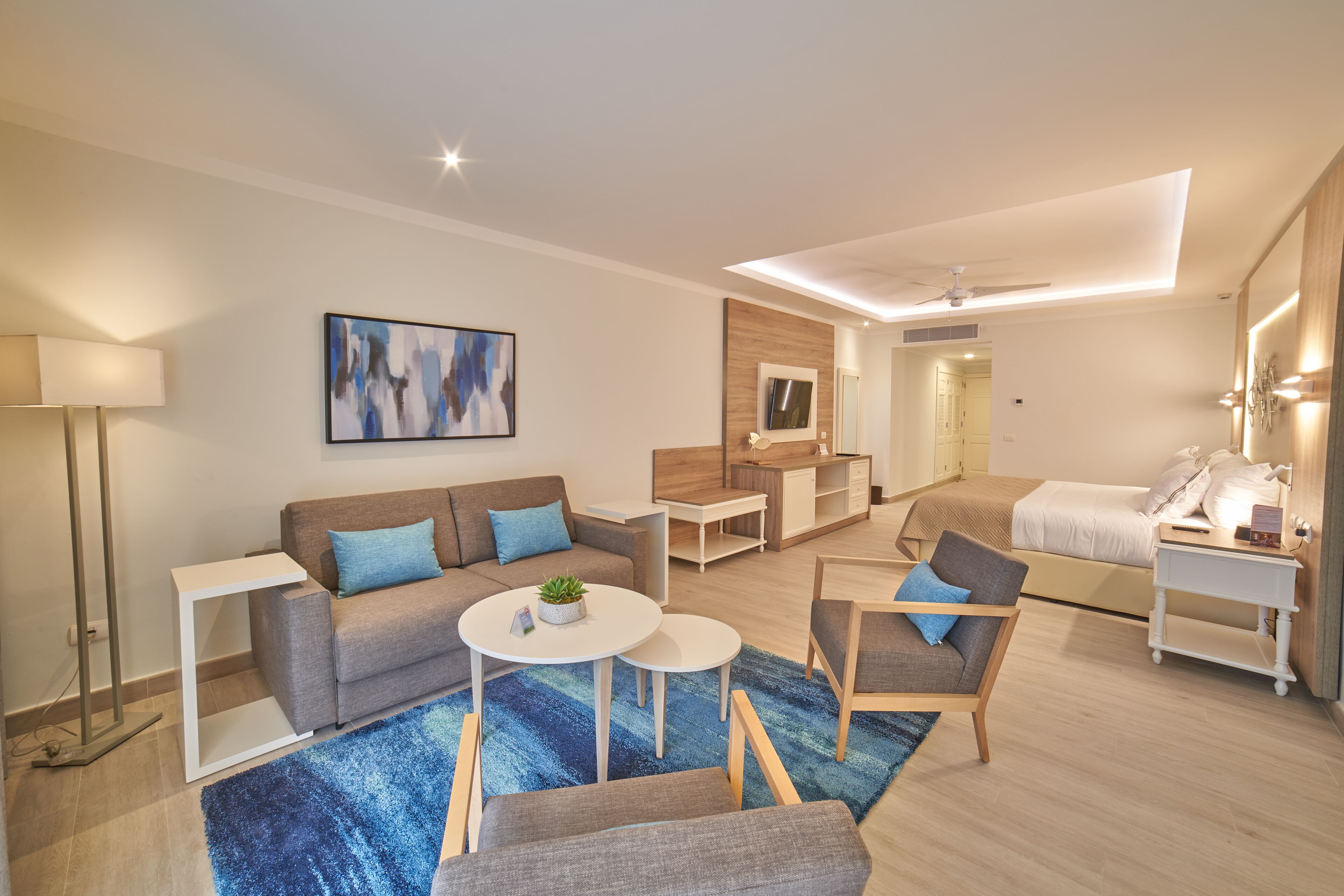 A modern, well-lit living area with a sofa, armchairs, a coffee table, and a TV. There is a kitchenette in the background and a separate bedroom area.