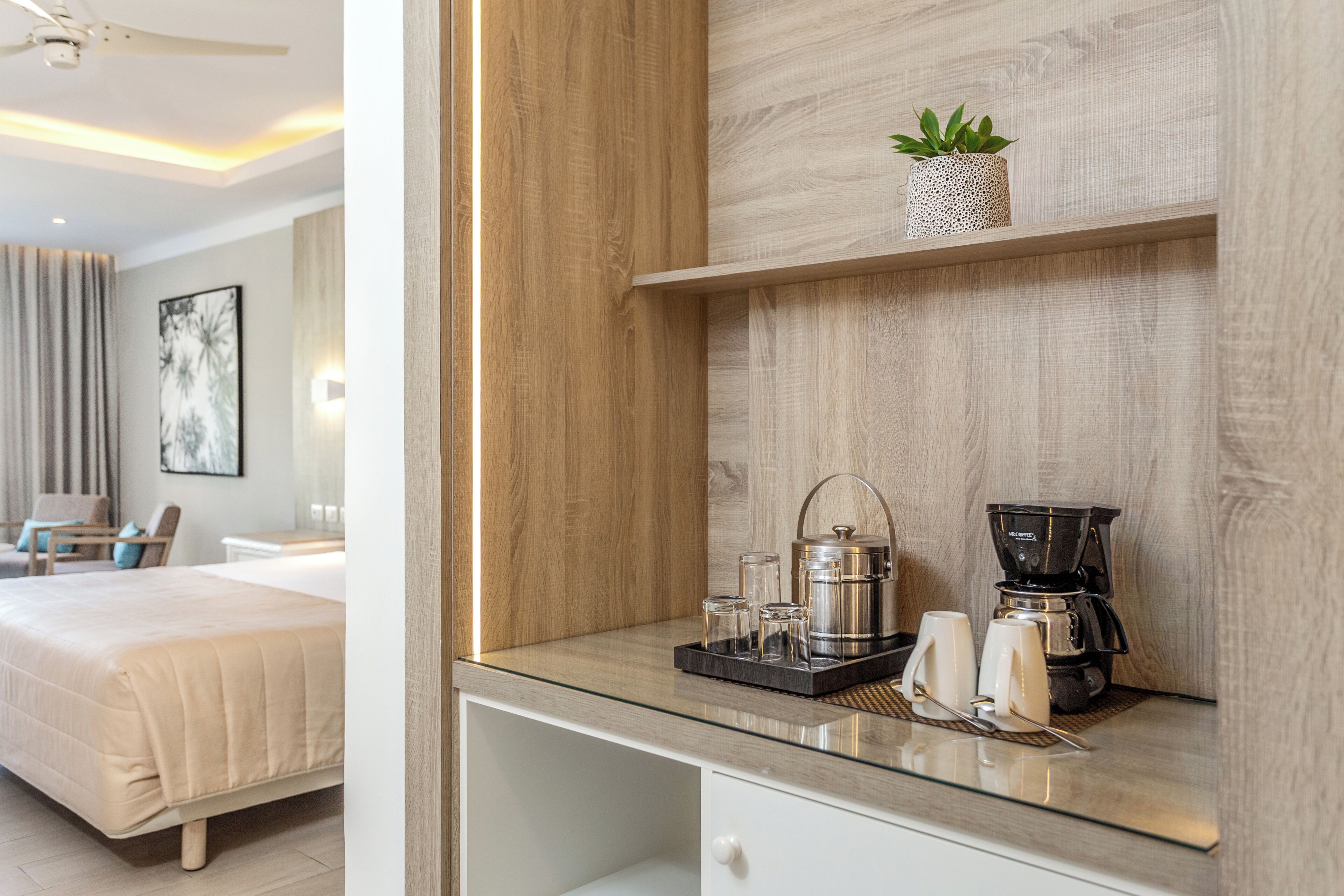 Une chambre d’hôtel avec un lit, une table de chevet sur laquelle se trouve une machine à café, et une petite plante en pot posée sur une étagère.