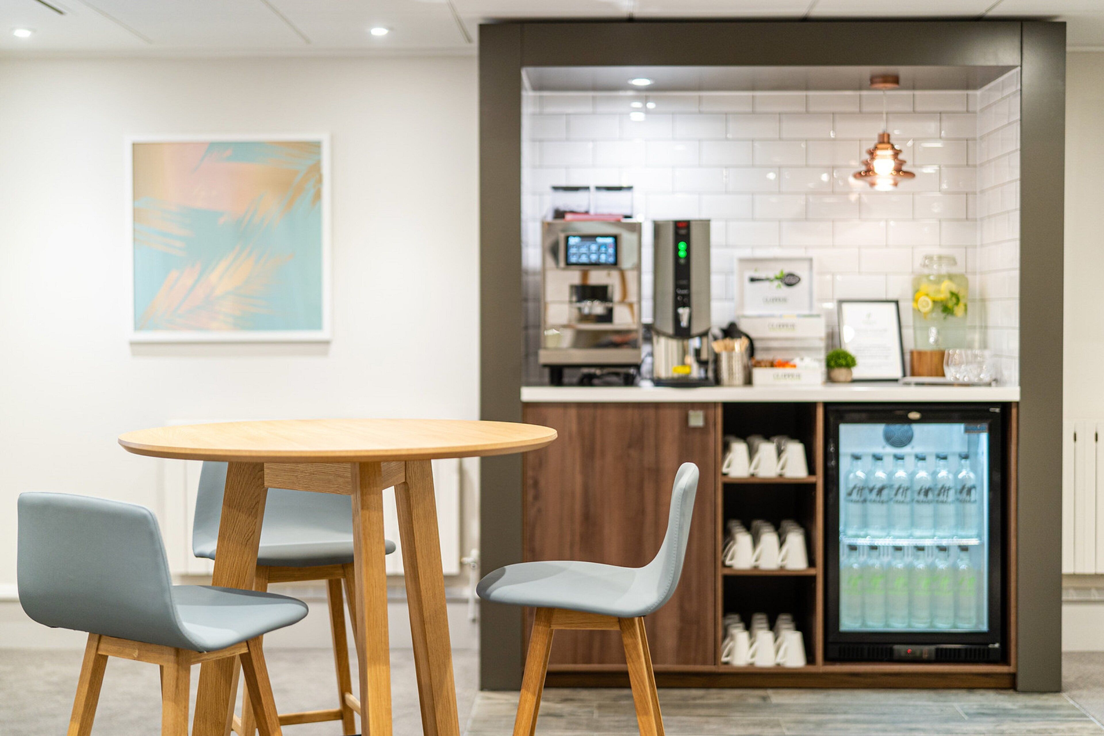 Un área de cocina moderna con una mesa redonda de madera y dos sillas, un refrigerador lleno de bebidas y una cafetera.