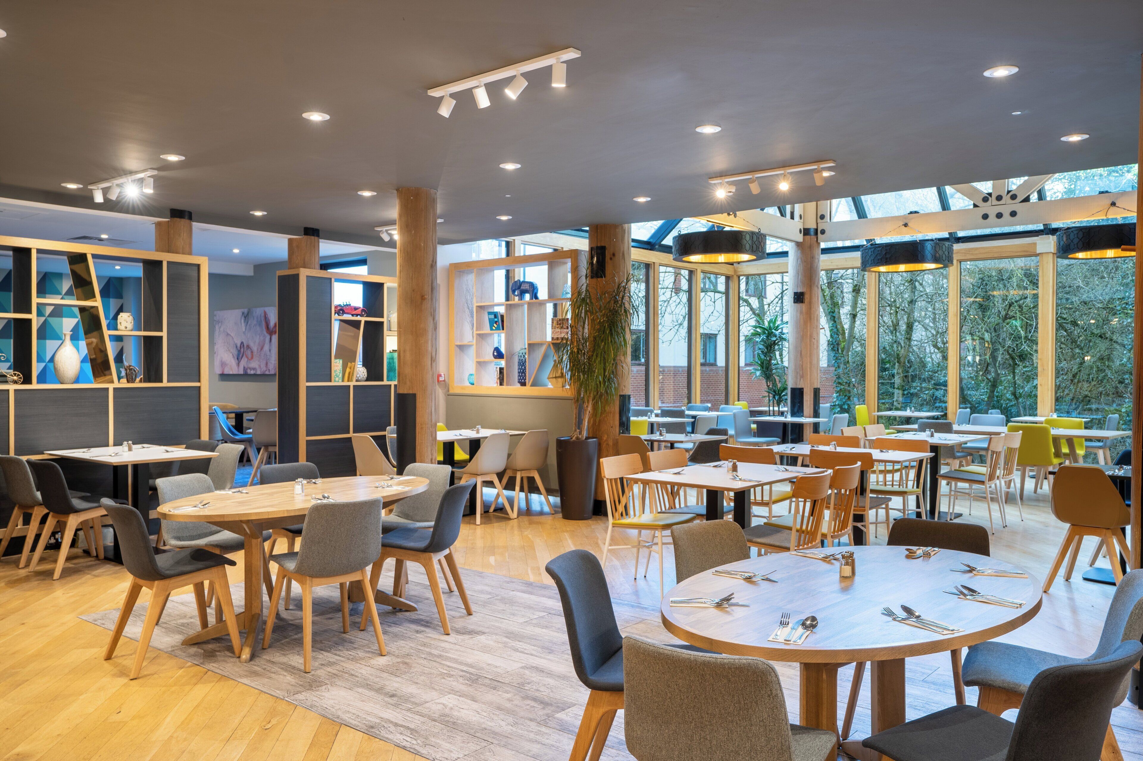 A modern dining area with round tables and chairs, wooden beams, and large windows offering views of the outdoors.
