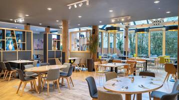 A modern dining area with round tables and chairs, wooden beams, and large windows offering views of the outdoors.