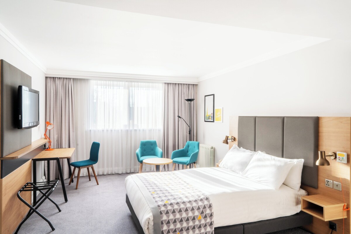 A hotel room with a bed, two chairs, a desk, a TV, and a window with curtains.