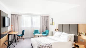 A hotel room with a bed, two chairs, a desk, a TV, and a window with curtains.