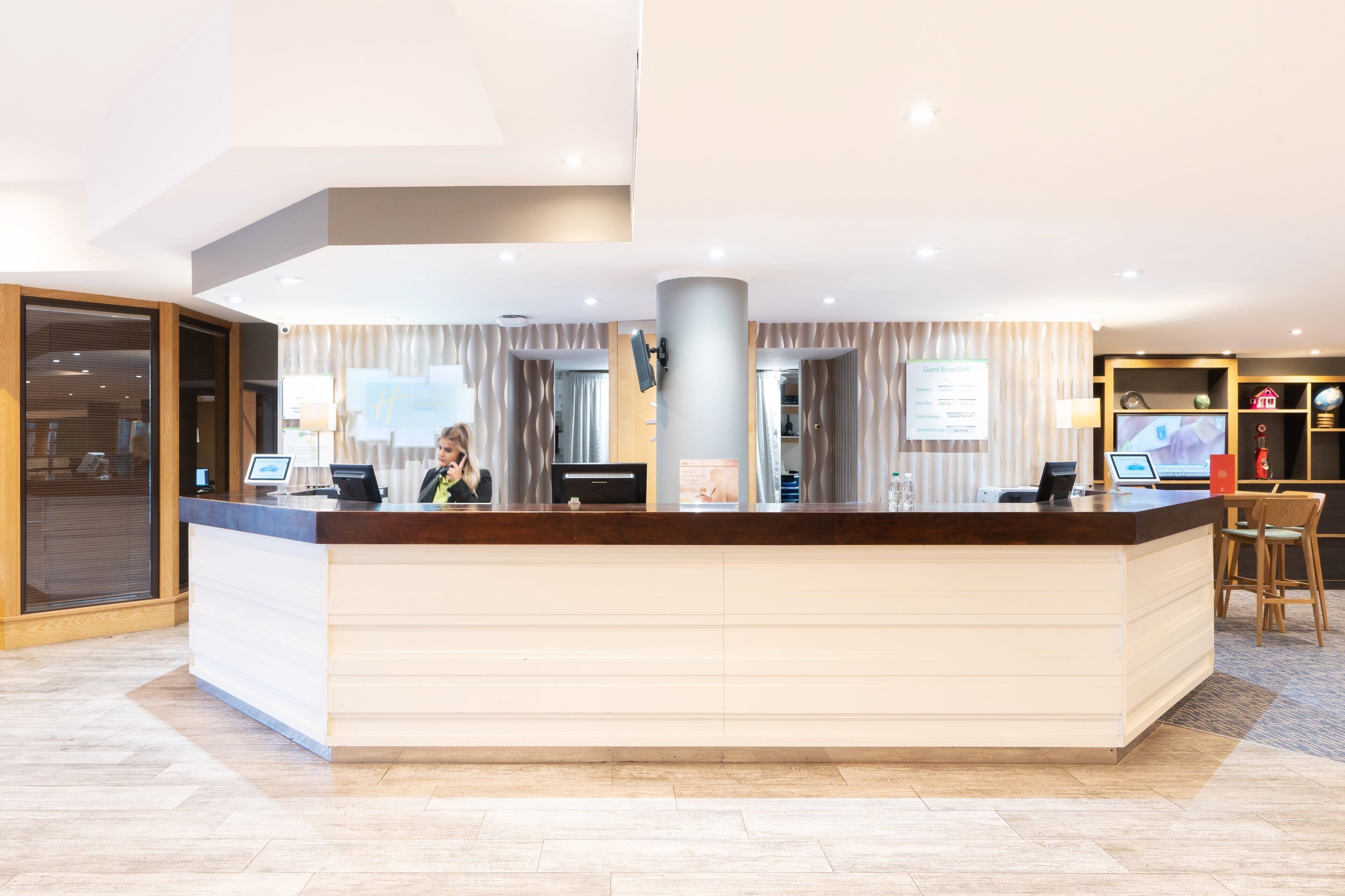 A hotel reception desk with a digital screen, a guest room tariff sign, and a reception area with a large window.