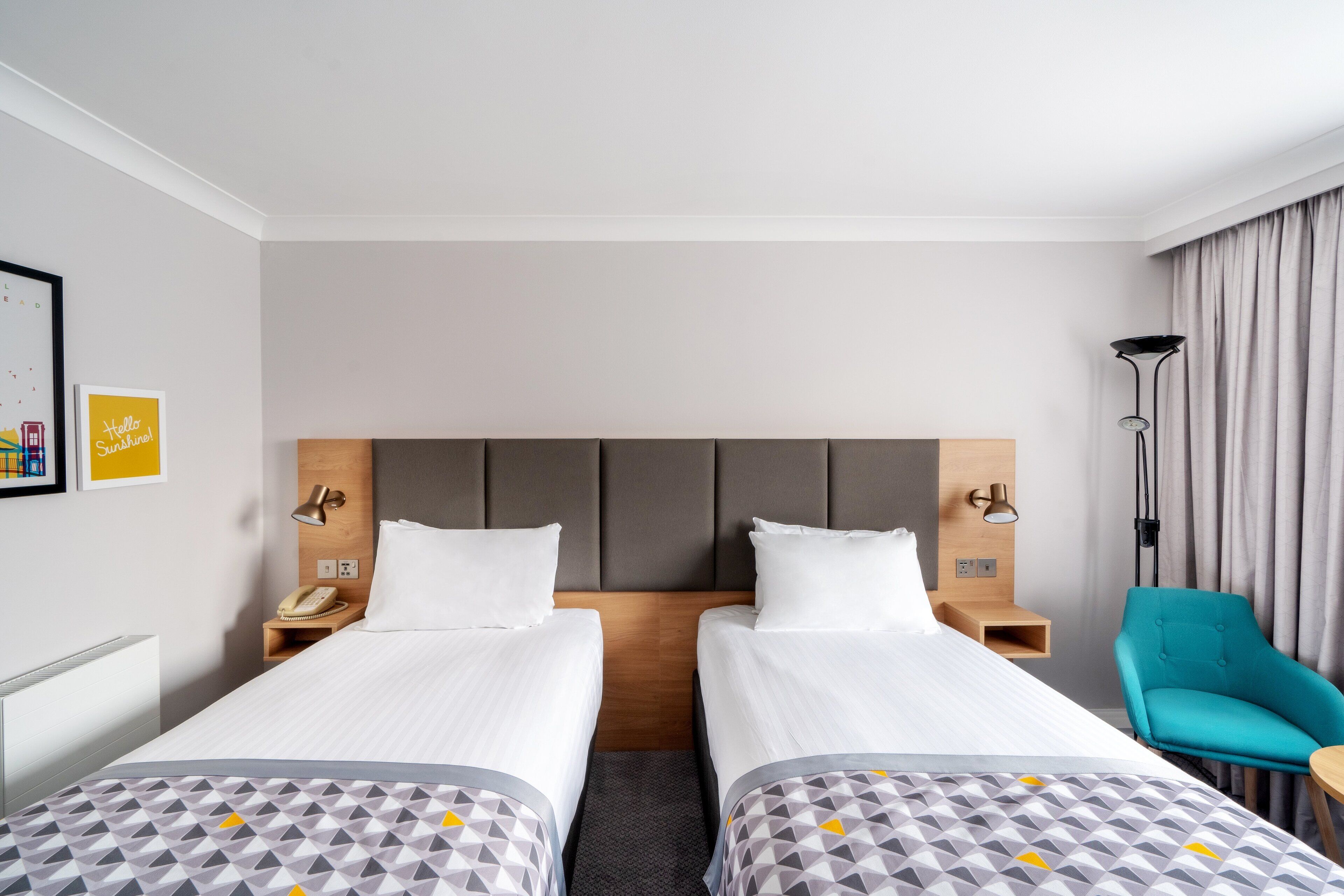 A hotel room with two single beds, a wooden headboard, bedside lamps, a teal armchair, and a small table.