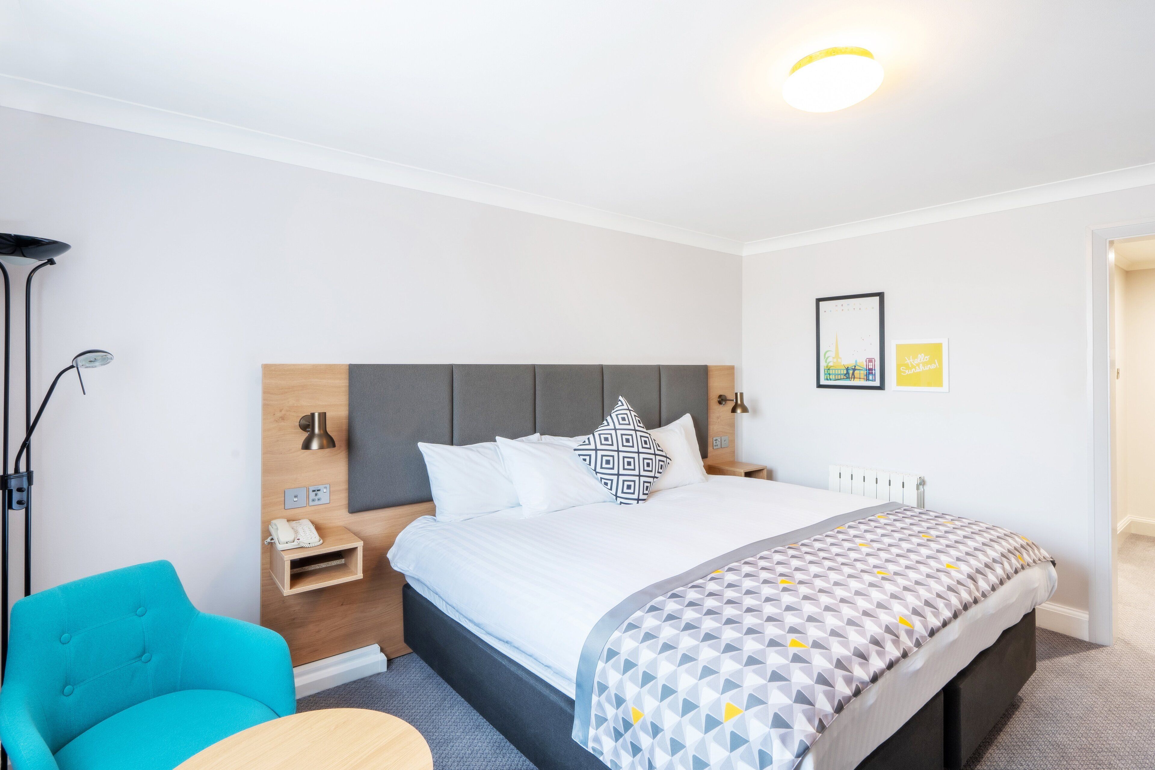 A modern hotel room with a large bed, a bedside table with a lamp, a chair, and a small desk.