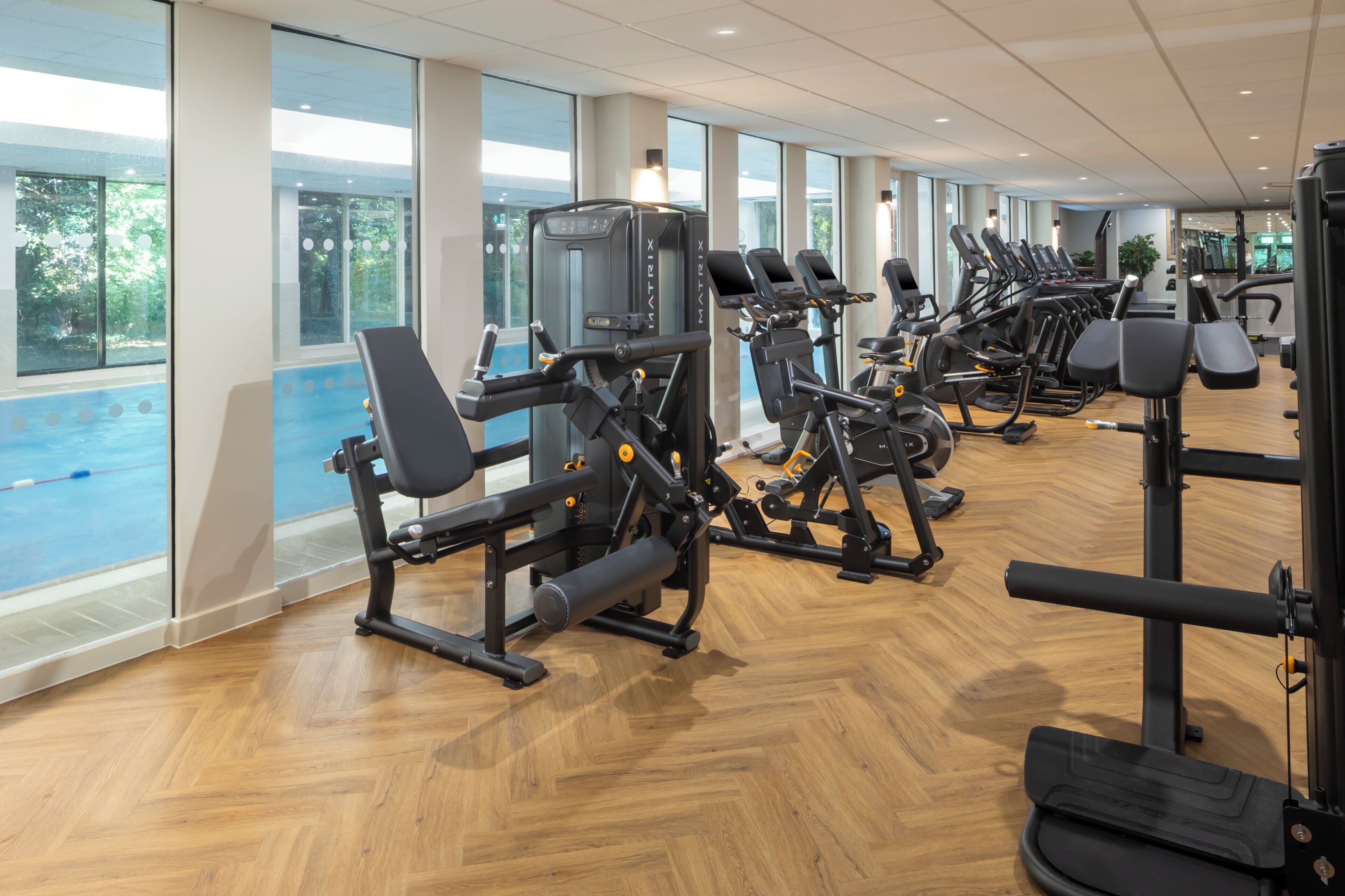 A gym with various exercise machines, large windows, and a view of an outdoor swimming pool.