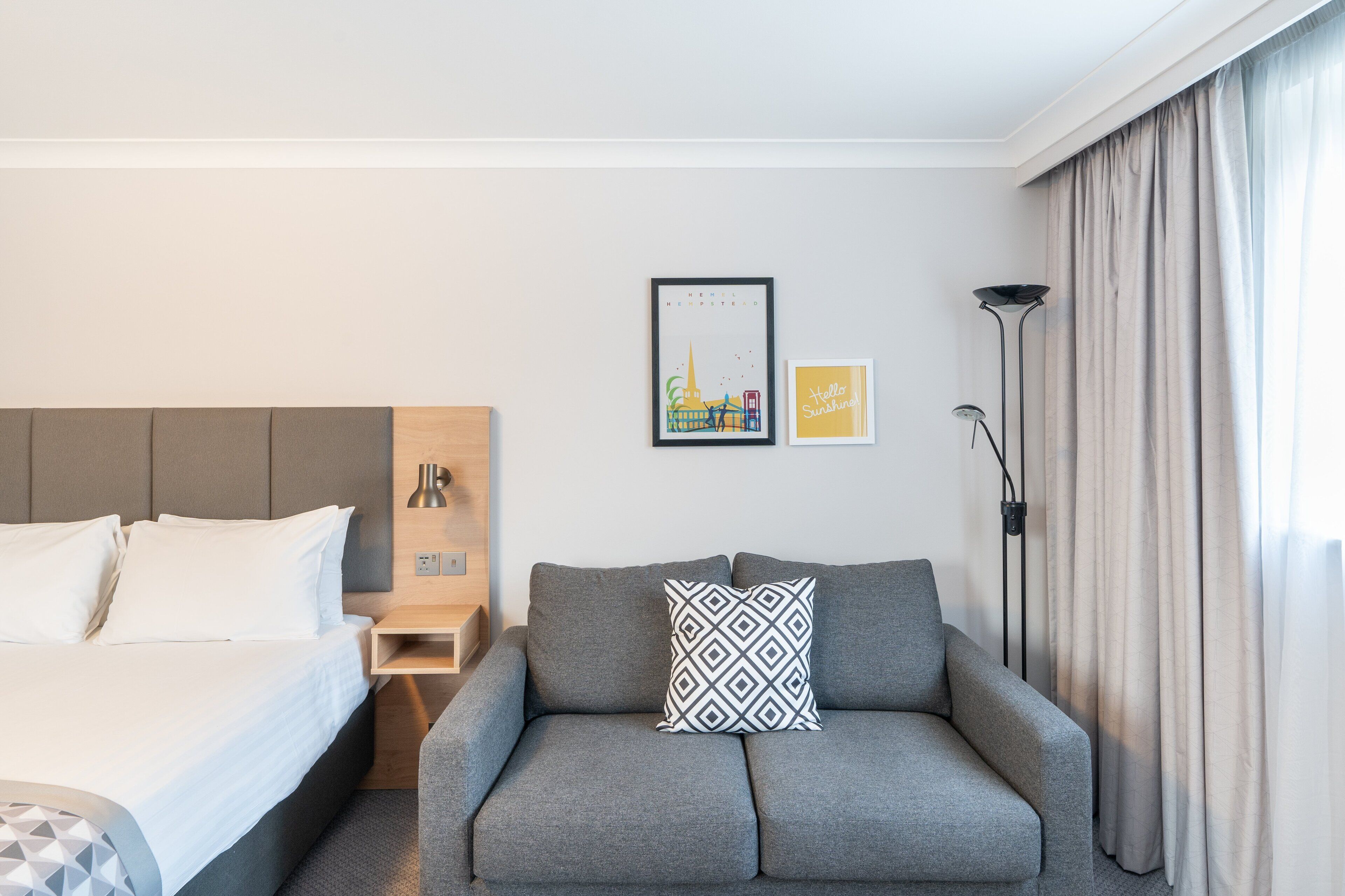 Quarto de hotel com uma cama, um sofá cinzento, uma mesa pequena e duas quadros na parede.