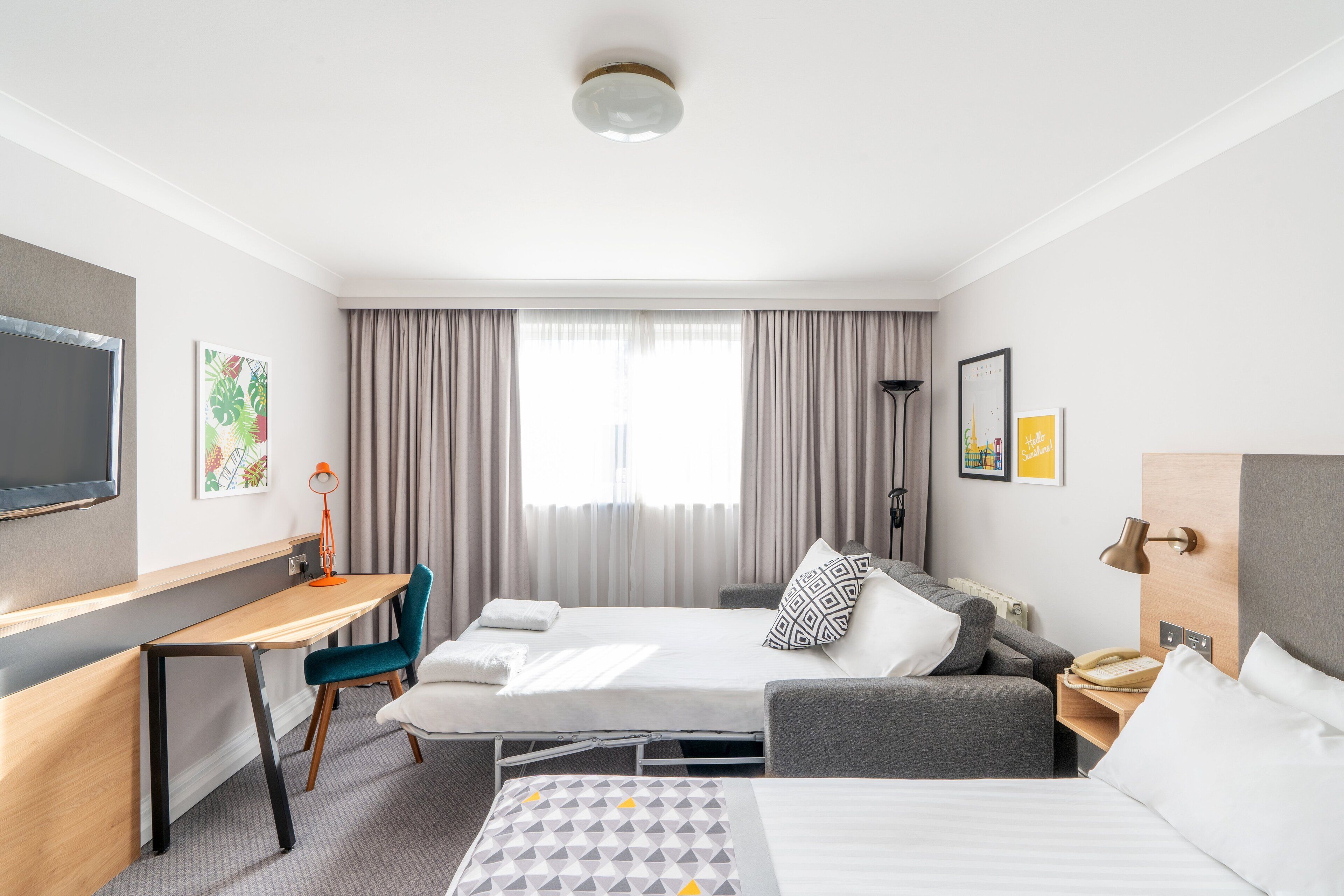 A hotel room with a bed, a desk, a chair, a sofa, a TV, and a window with curtains.