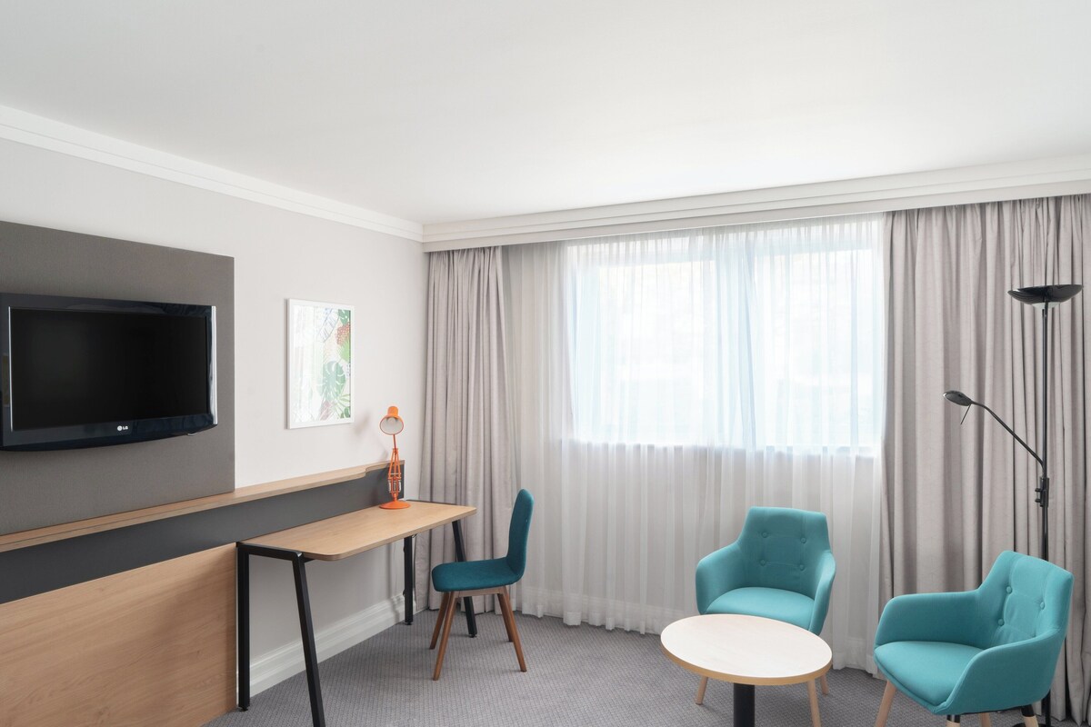 A modern hotel room with a flat-screen TV mounted on the wall, a wooden desk, two teal chairs, a round table, and a large window with sheer curtains.