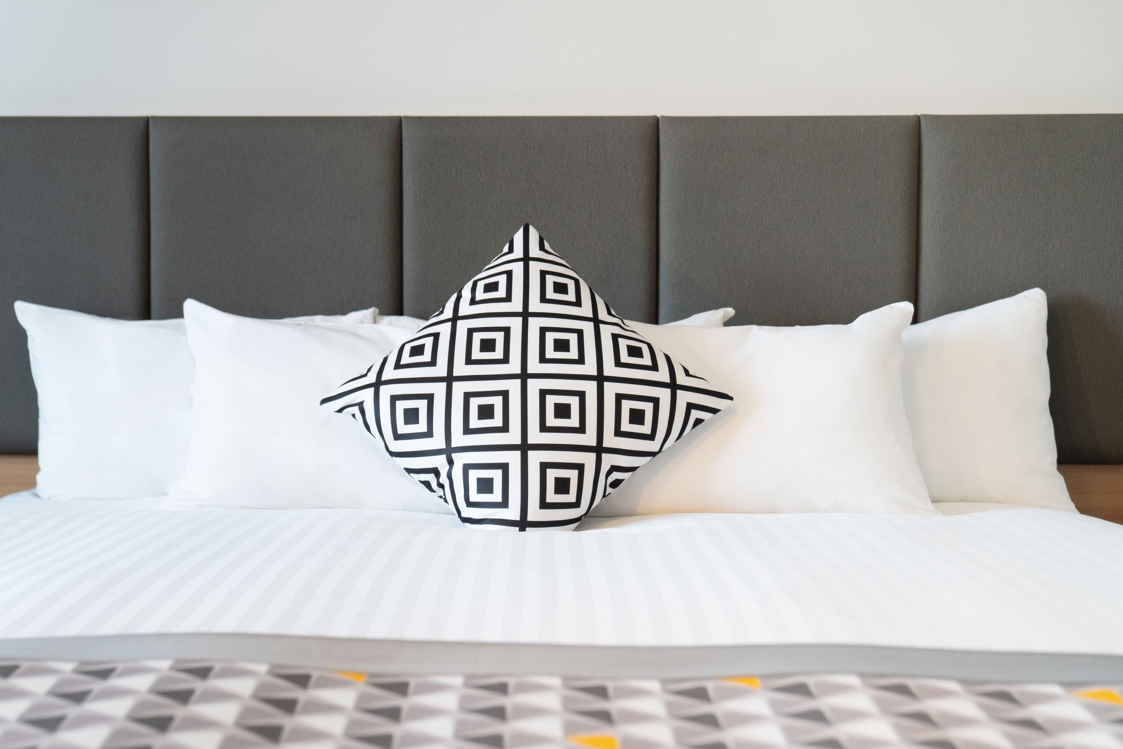 A bed with a grey upholstered headboard, white bedding, and a decorative pillow with a geometric pattern.