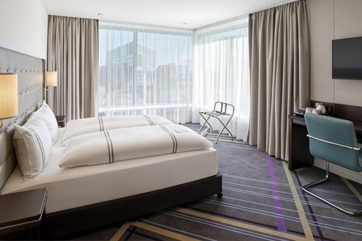A hotel room with a large bed, a desk, a chair, and a view of the city through large windows.