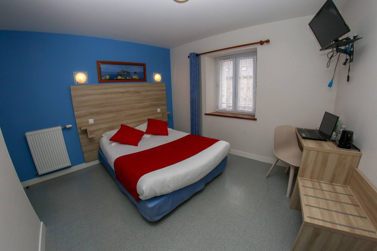 Ein Schlafzimmer mit Bett, Schreibtisch, Stuhl, Heizkörper, einem Fenster mit Vorhängen und einem an der Wand befestigten Fernseher.
