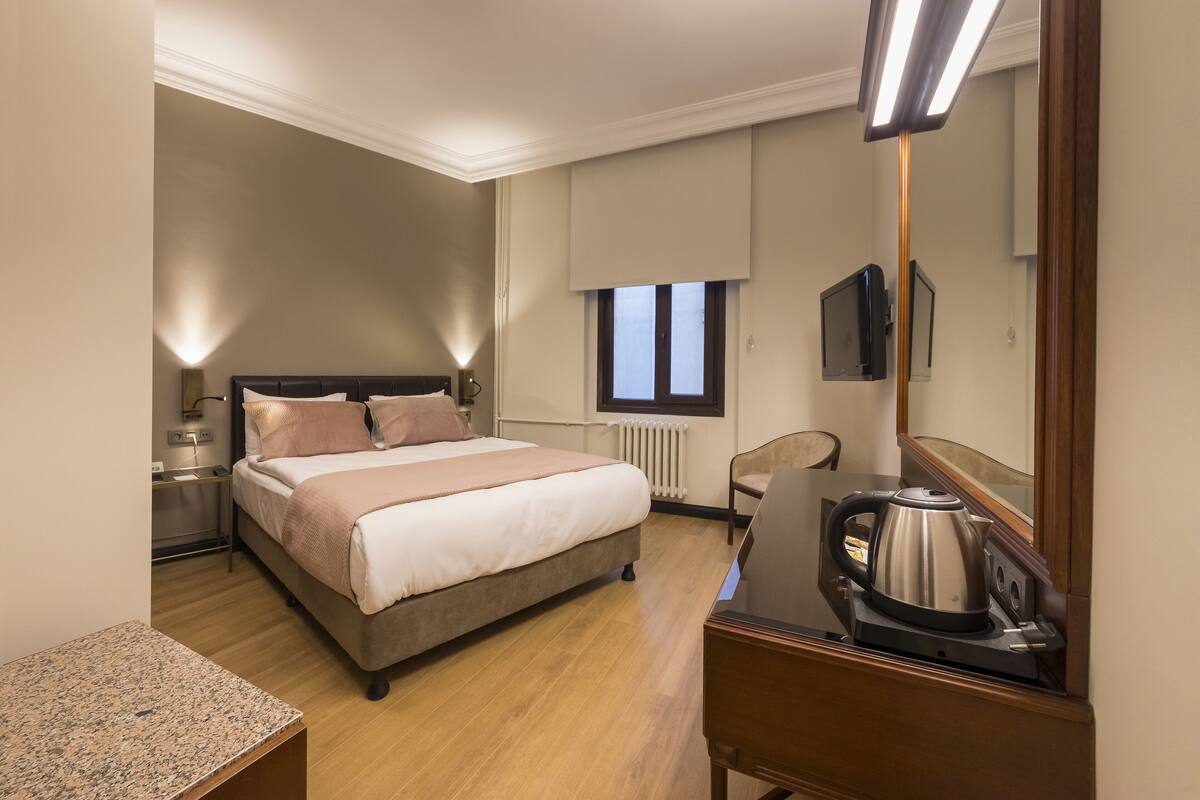 A hotel room with a bed, bedside table, lamp, chair, and a kettle on a table.