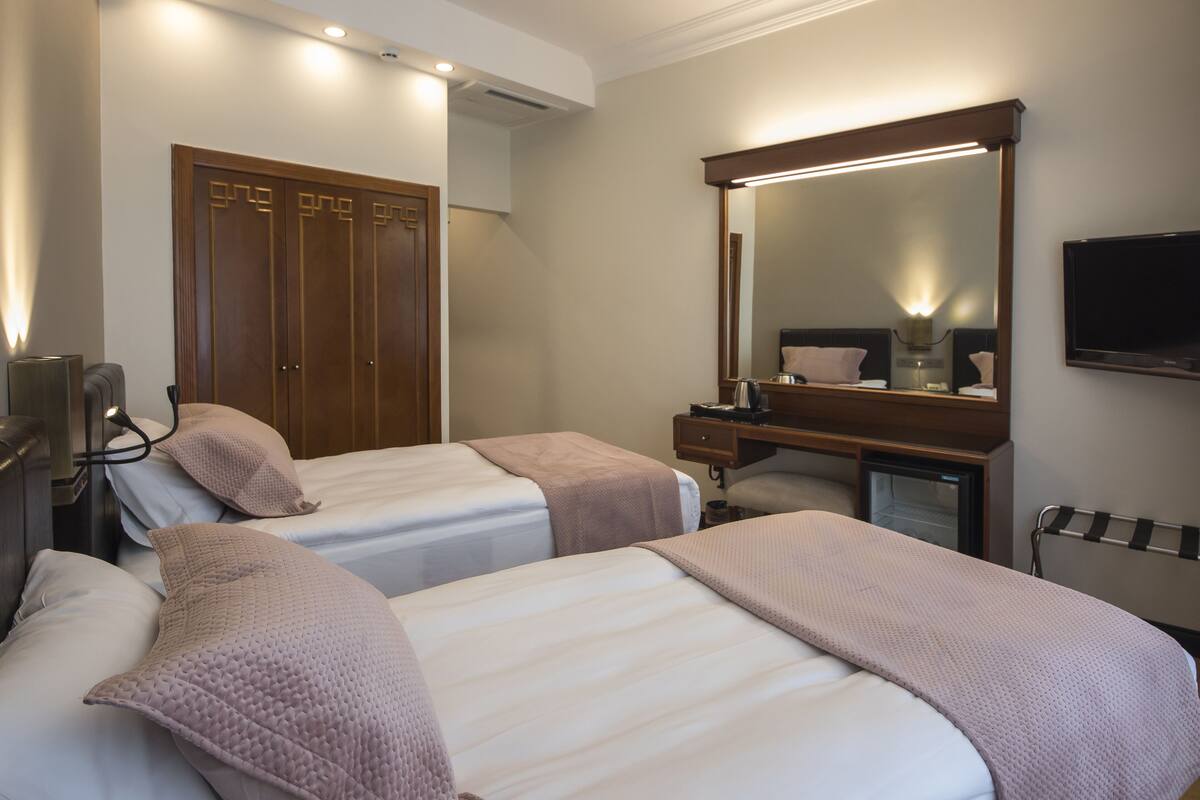 A hotel room with two beds, a wooden wardrobe, a mirror, a desk, and a television.