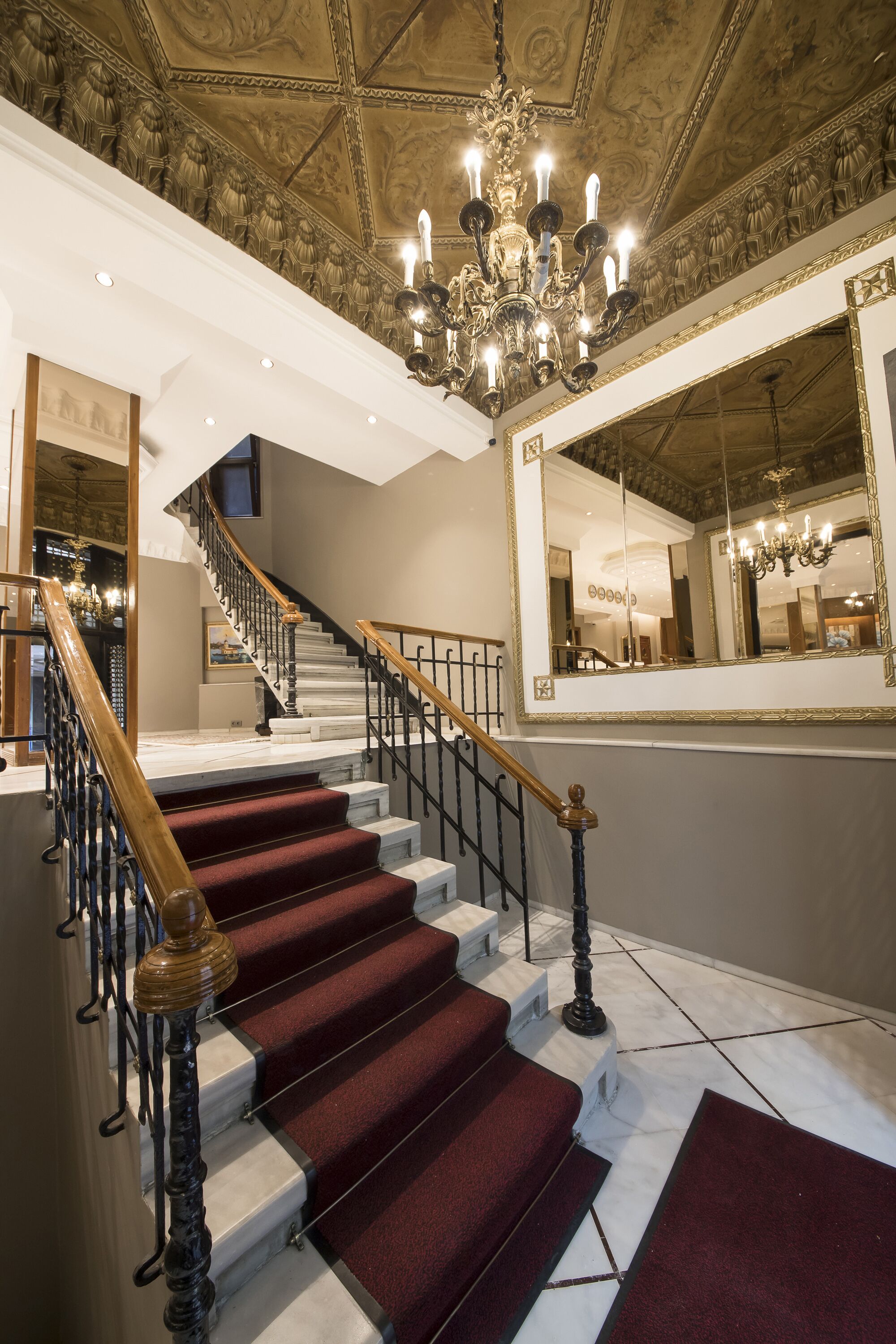 A grand staircase with red carpeted steps and a wooden handrail, leading to a landing with a large mirror and a chandelier.