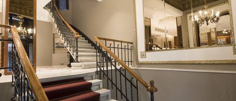 A grand staircase with red carpeted steps and a wooden handrail, leading to a landing with a large mirror and a chandelier.