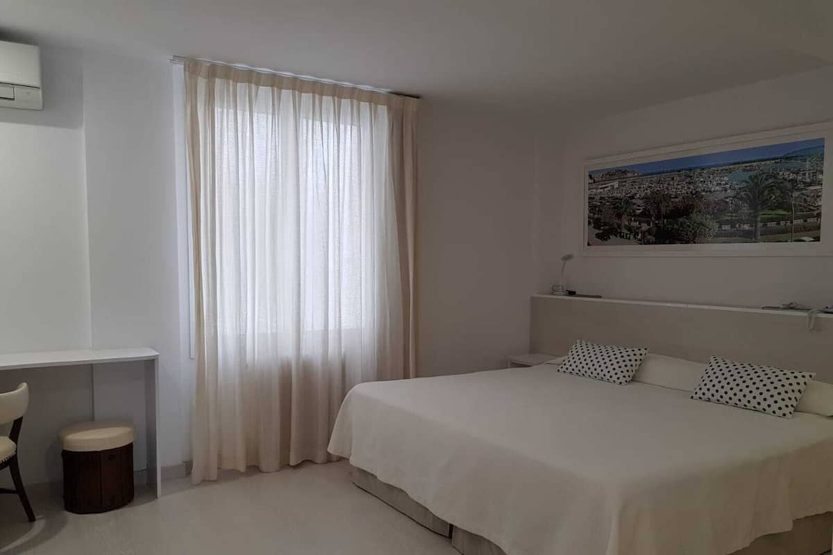 A bedroom with a bed, a desk, a chair, a small stool, and a window with sheer curtains.