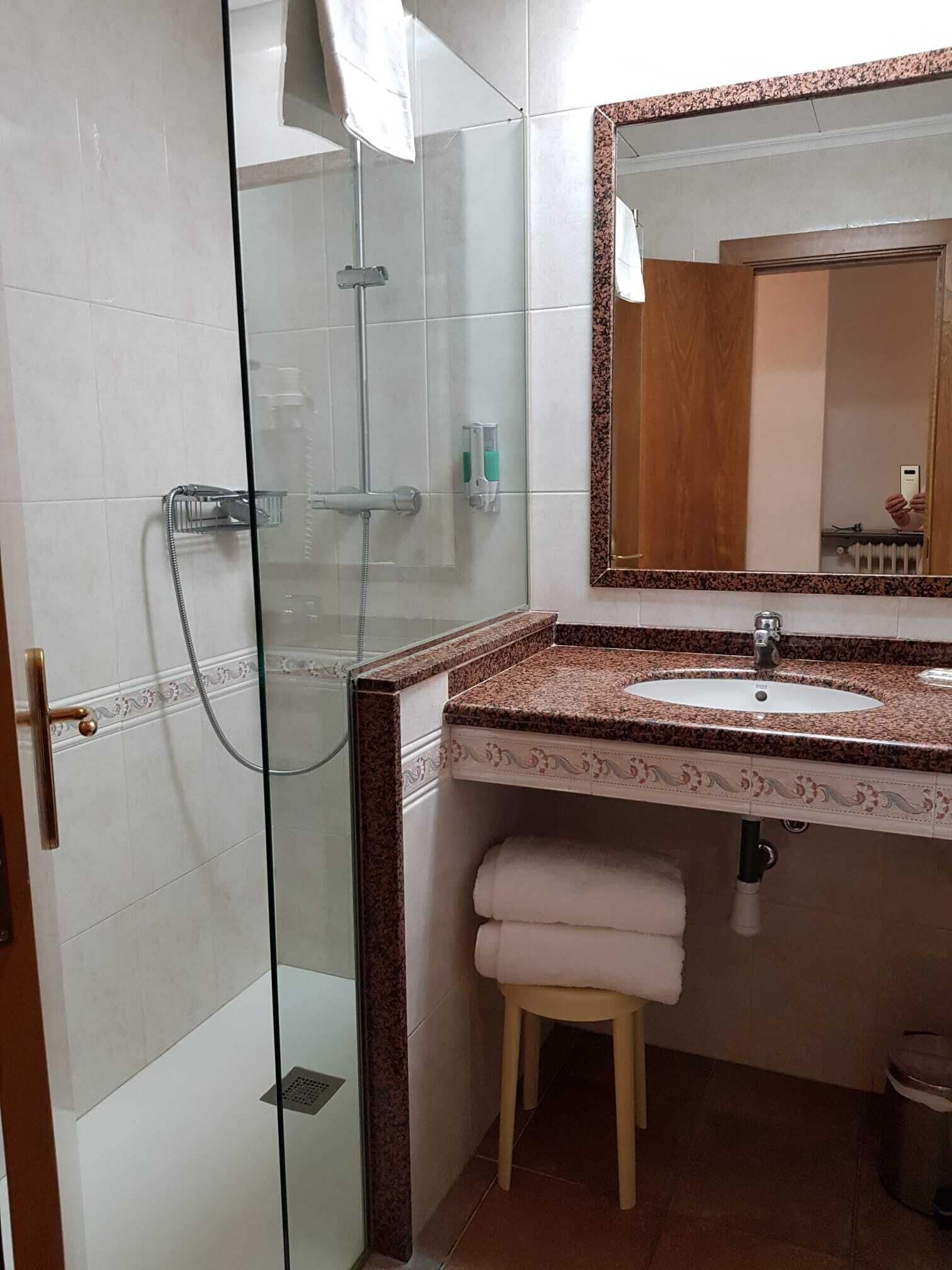 A bathroom with a glass shower enclosure, a sink with a marble countertop, a wooden stool, and a mirror with a wooden frame.
