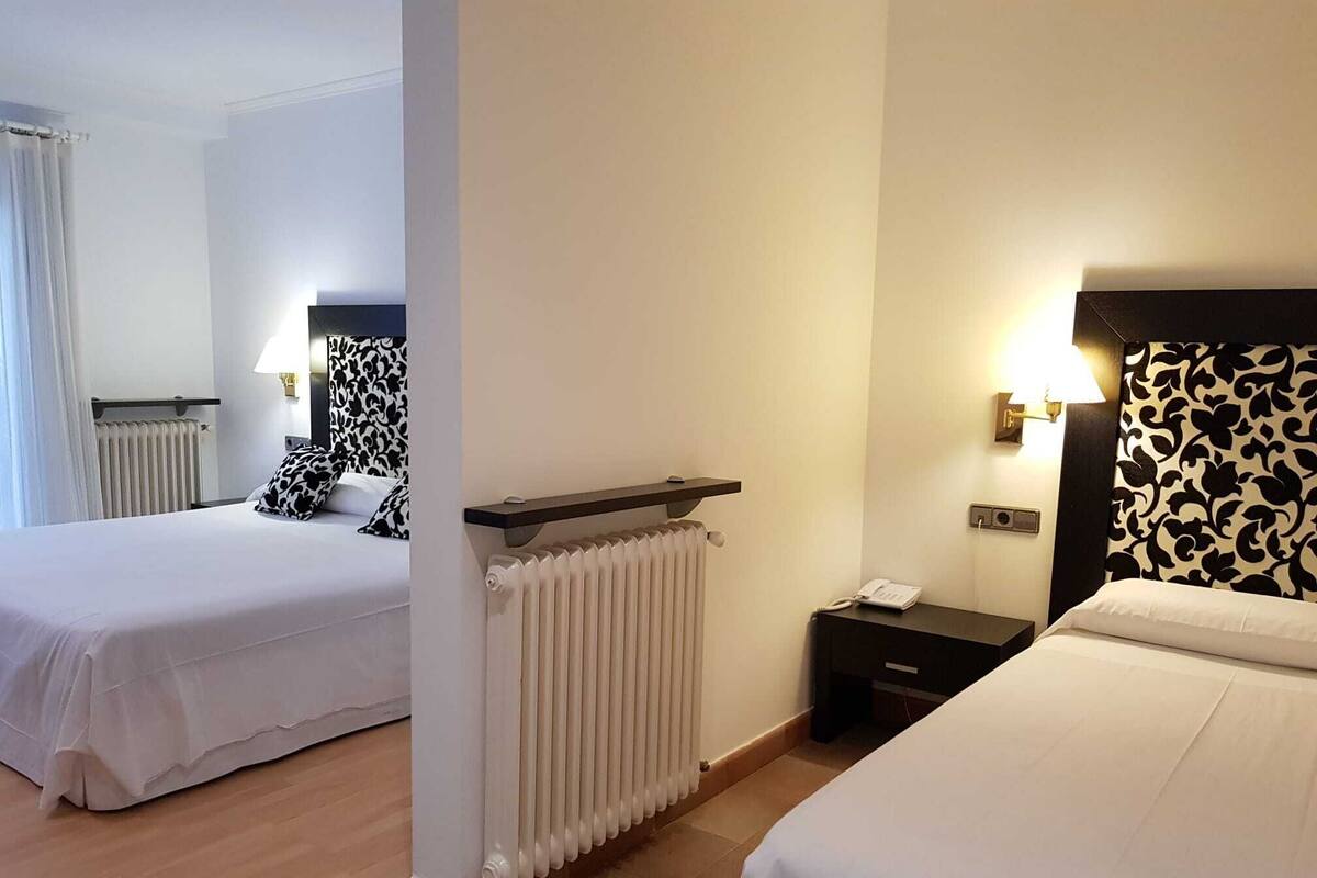 A hotel room with two beds, a radiator, a nightstand with a lamp, and a telephone.