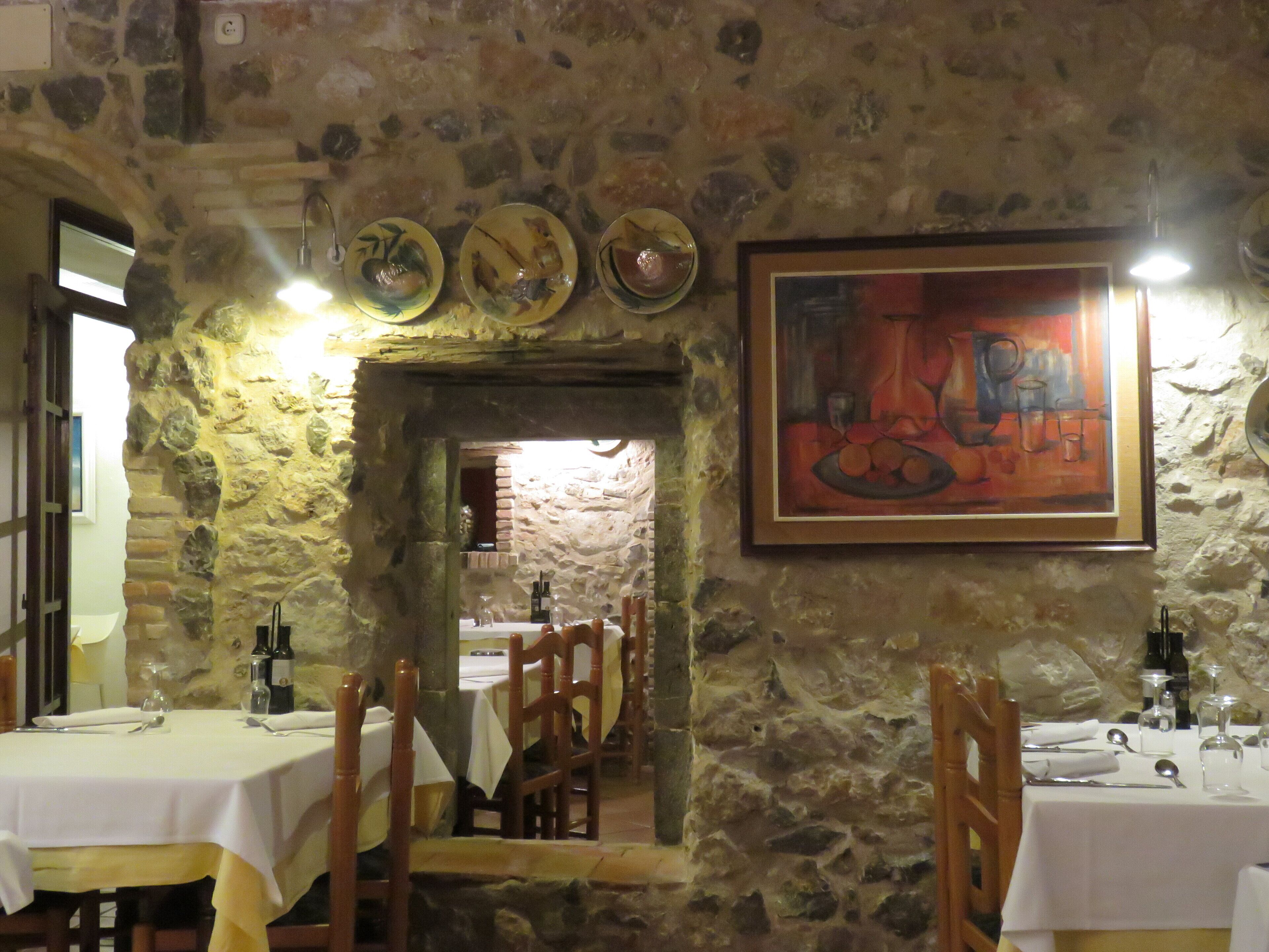 A rustic dining area with stone walls, wooden chairs, and tables set for dining. There is a framed painting on the wall and a doorway leading to another room.