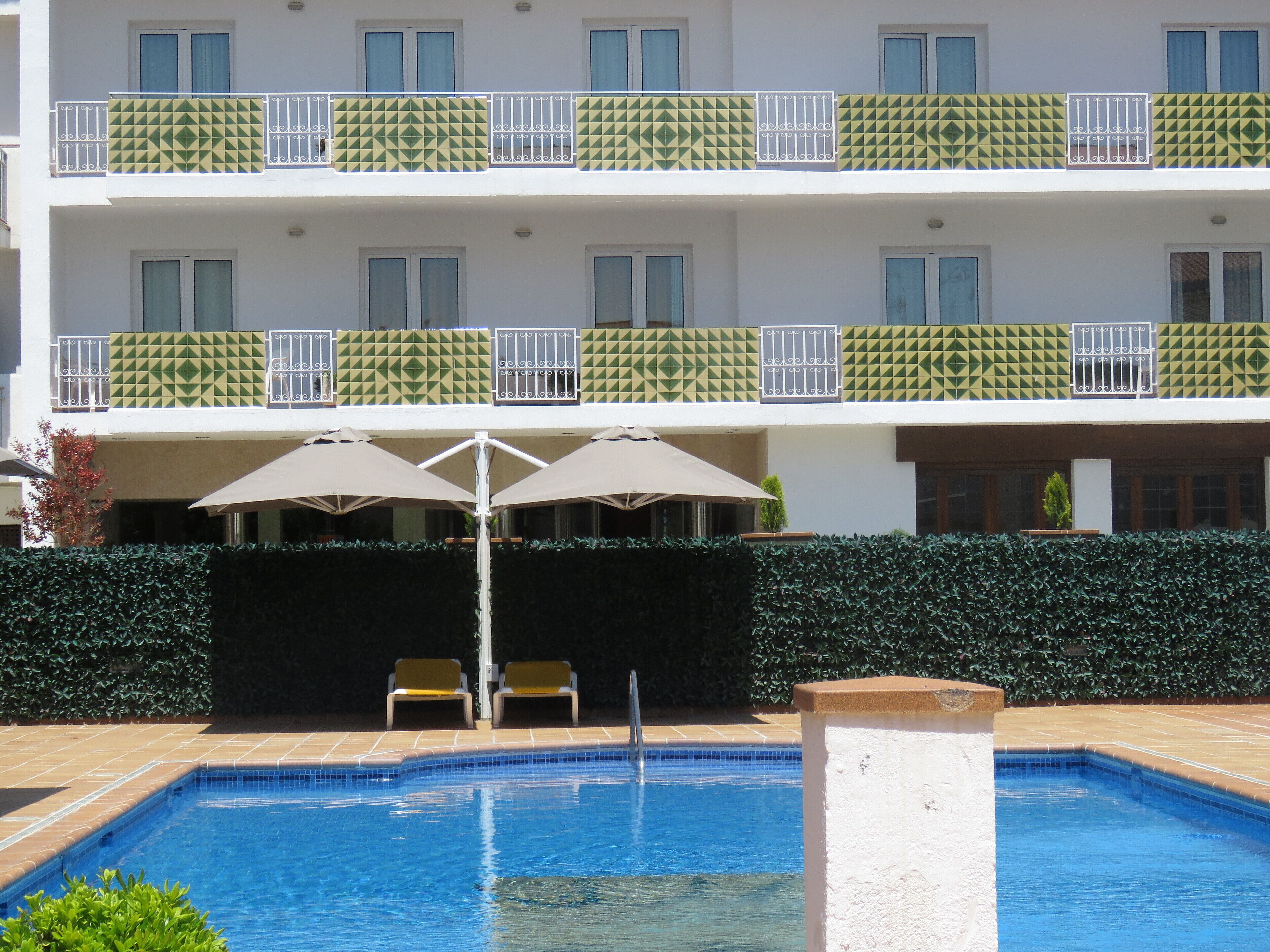 Una piscina con borde de azulejos, rodeada por un seto, y un edificio con balcones al fondo.