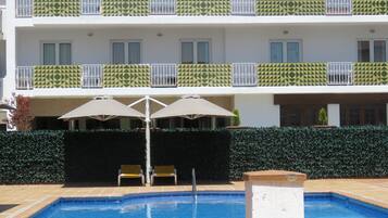 Una piscina con borde de azulejos, rodeada por un seto, y un edificio con balcones al fondo.