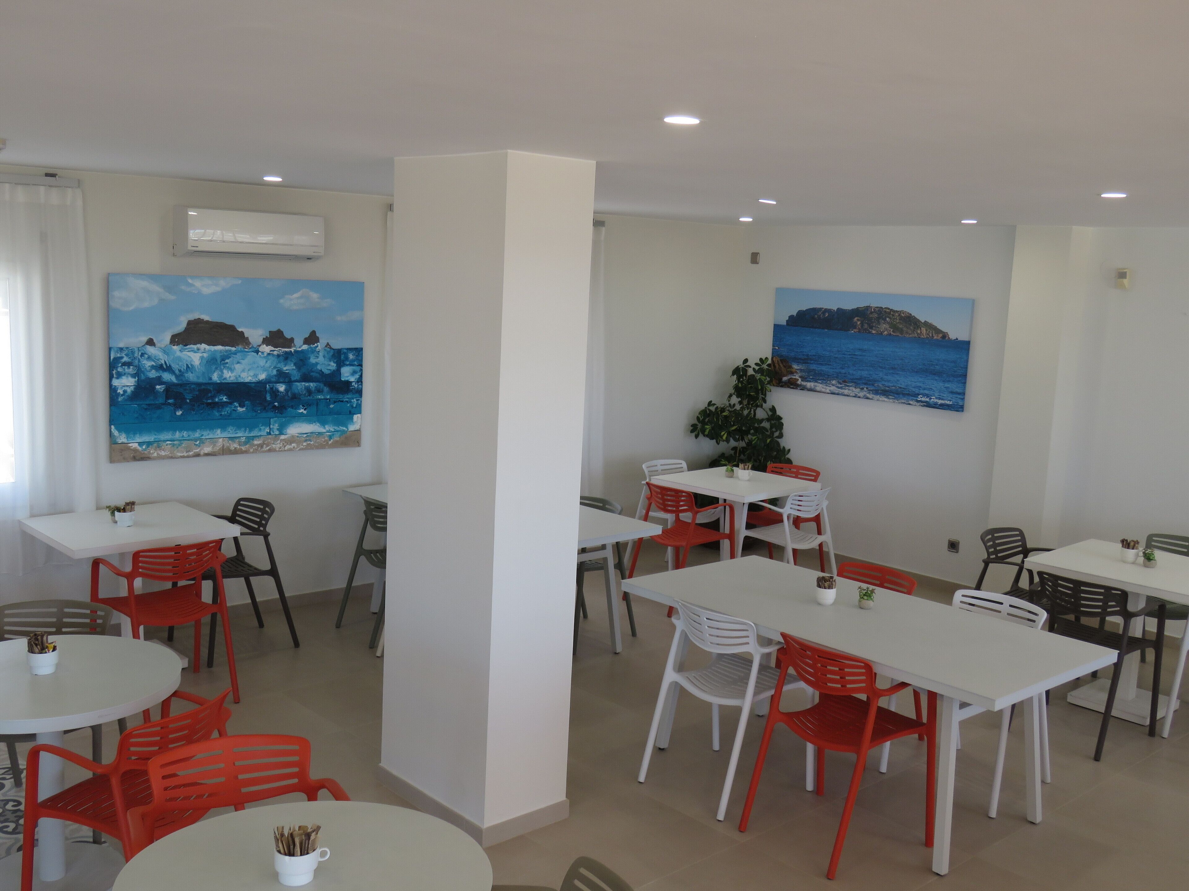 Un comedor con mesas blancas y sillas rojas, dos cuadros con paisajes costeros y un equipo de aire acondicionado en la pared.
