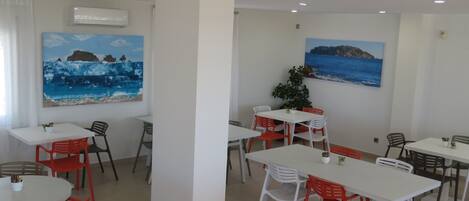 Un comedor con mesas blancas y sillas rojas, dos cuadros con paisajes costeros y un equipo de aire acondicionado en la pared.